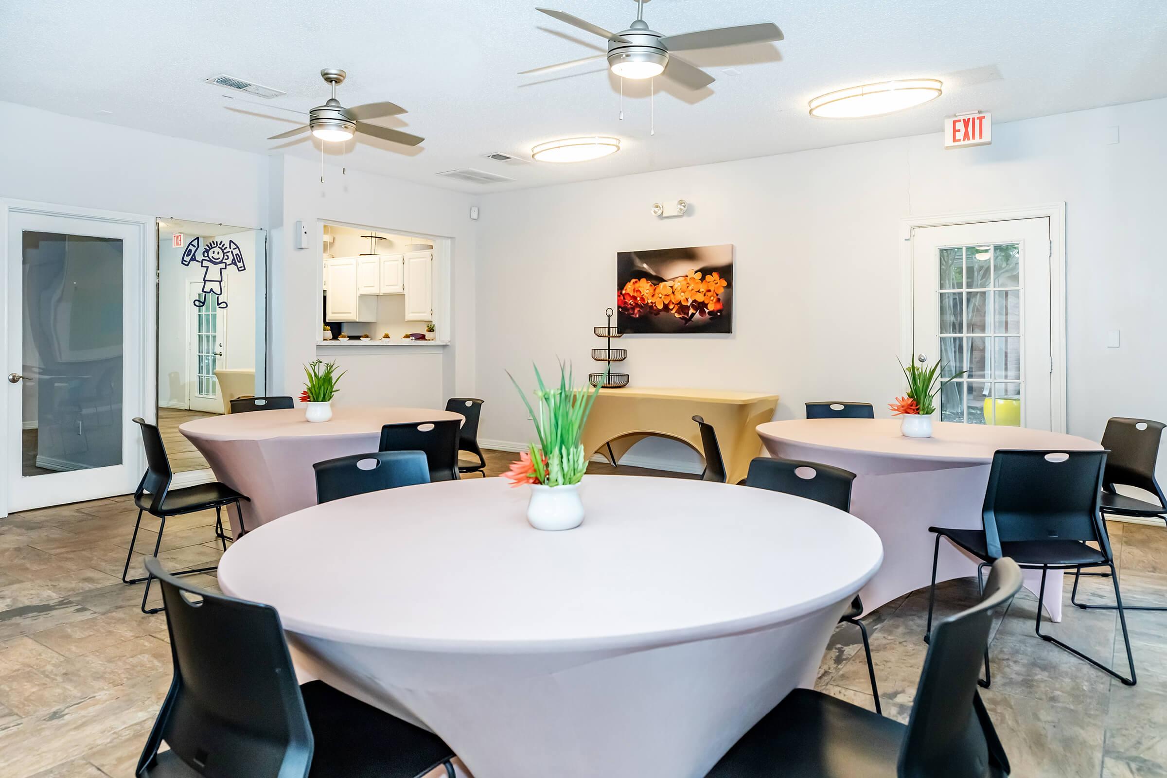 A bright, spacious gathering room featuring several round tables with light beige table covers. Each table has a small potted plant in the center. The room has light-colored walls, ceiling fans, and a modern artwork piece on the wall. A doorway leads to a kitchen area in the background.