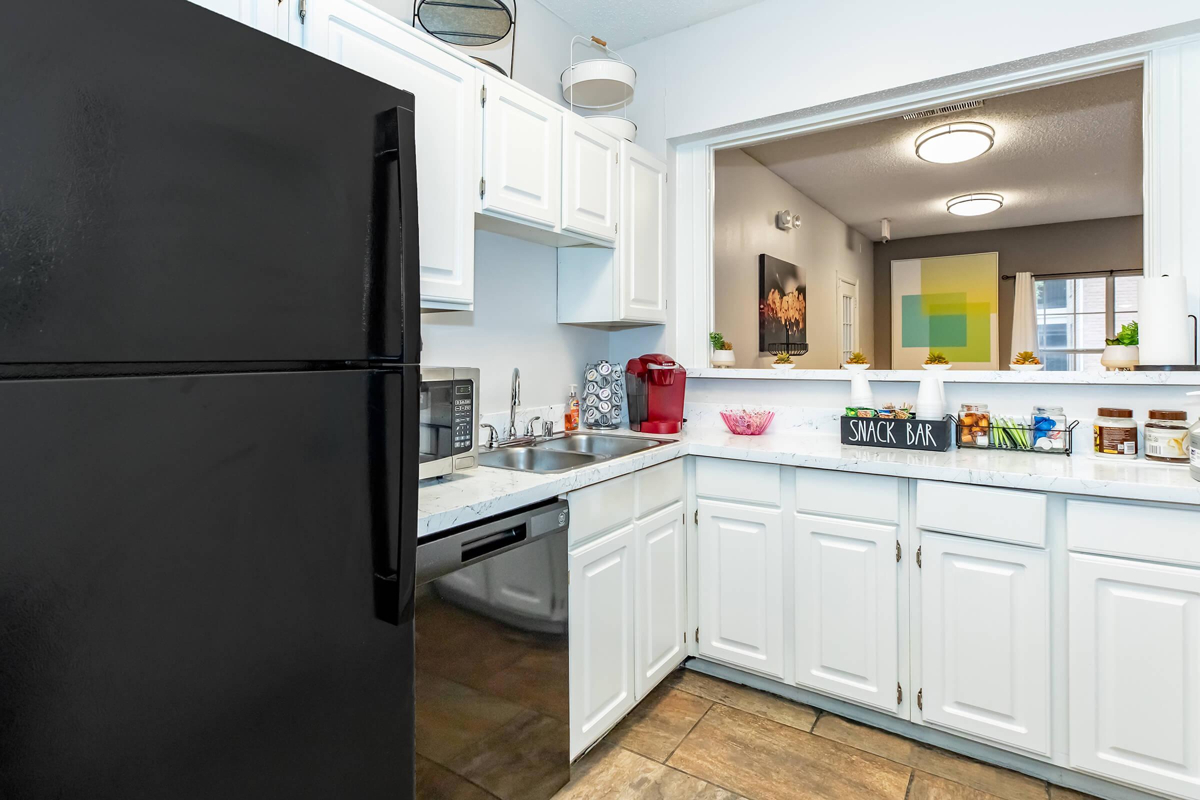 A modern kitchen featuring white cabinets, a black refrigerator, and stainless steel appliances. The countertop is decorated with a snack bar sign and various containers. A small window opens to a living space with colorful artwork, providing a bright and inviting atmosphere.