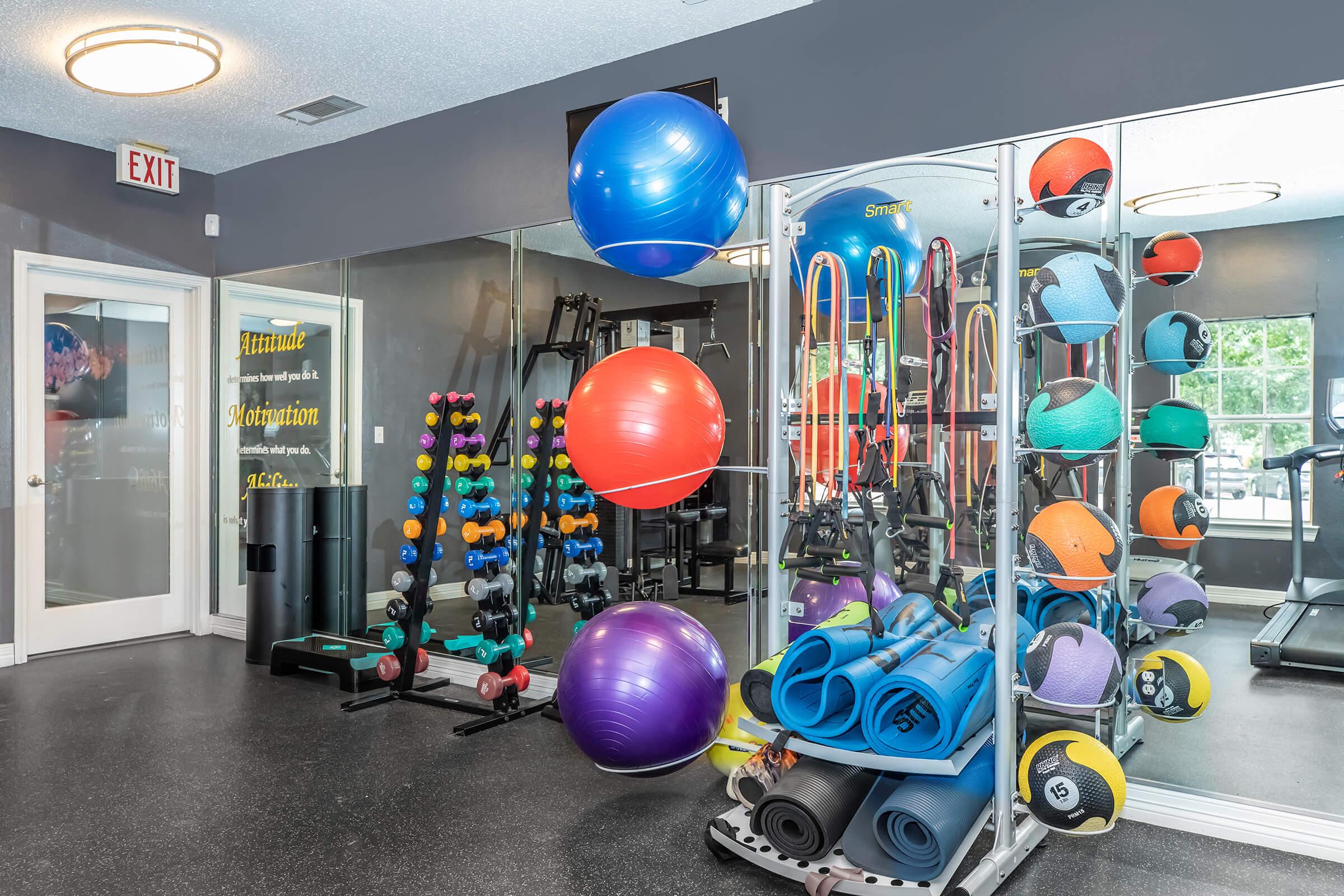 A well-equipped fitness area featuring various exercise equipment, including colorful exercise balls, dumbbells, yoga mats, and medicine balls. The walls are adorned with mirrors, reflecting the workout space, and there is a dedicated area for strength training.