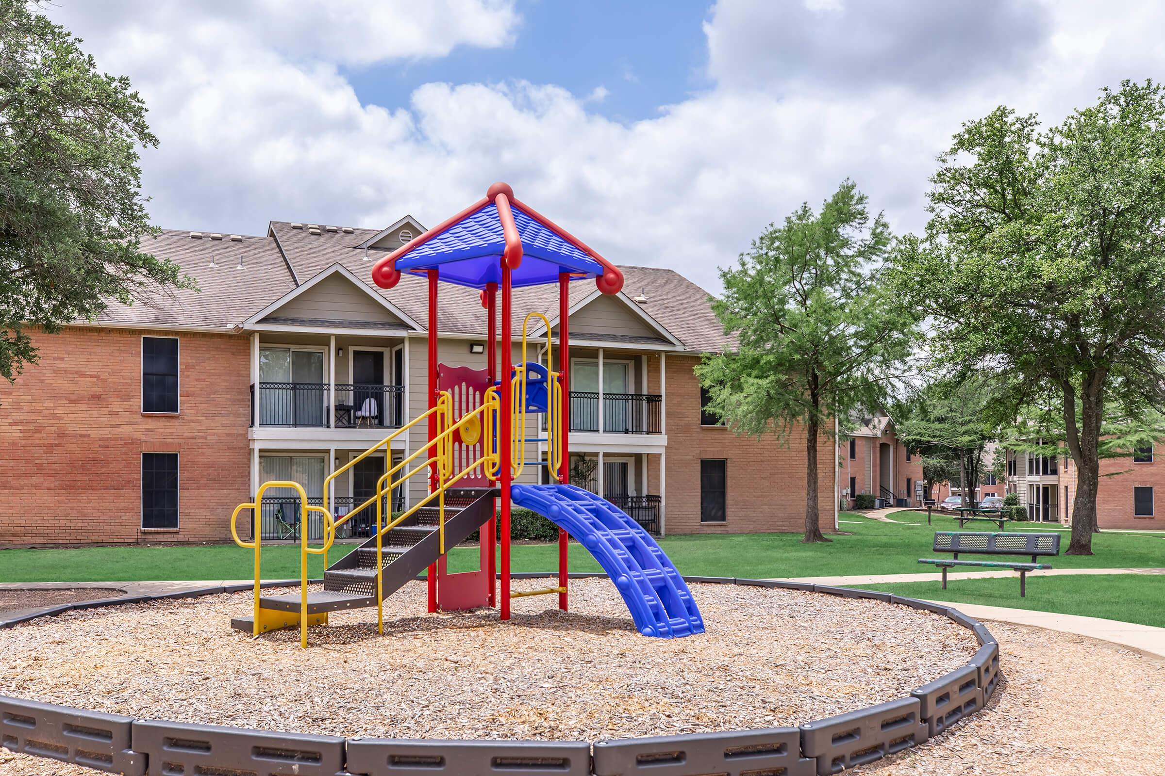 A colorful playground with a slide and climbing structure sits on a gravel surface in a grassy area, surrounded by brick apartment buildings. In the background, there are trees and a bench, under a partly cloudy sky.