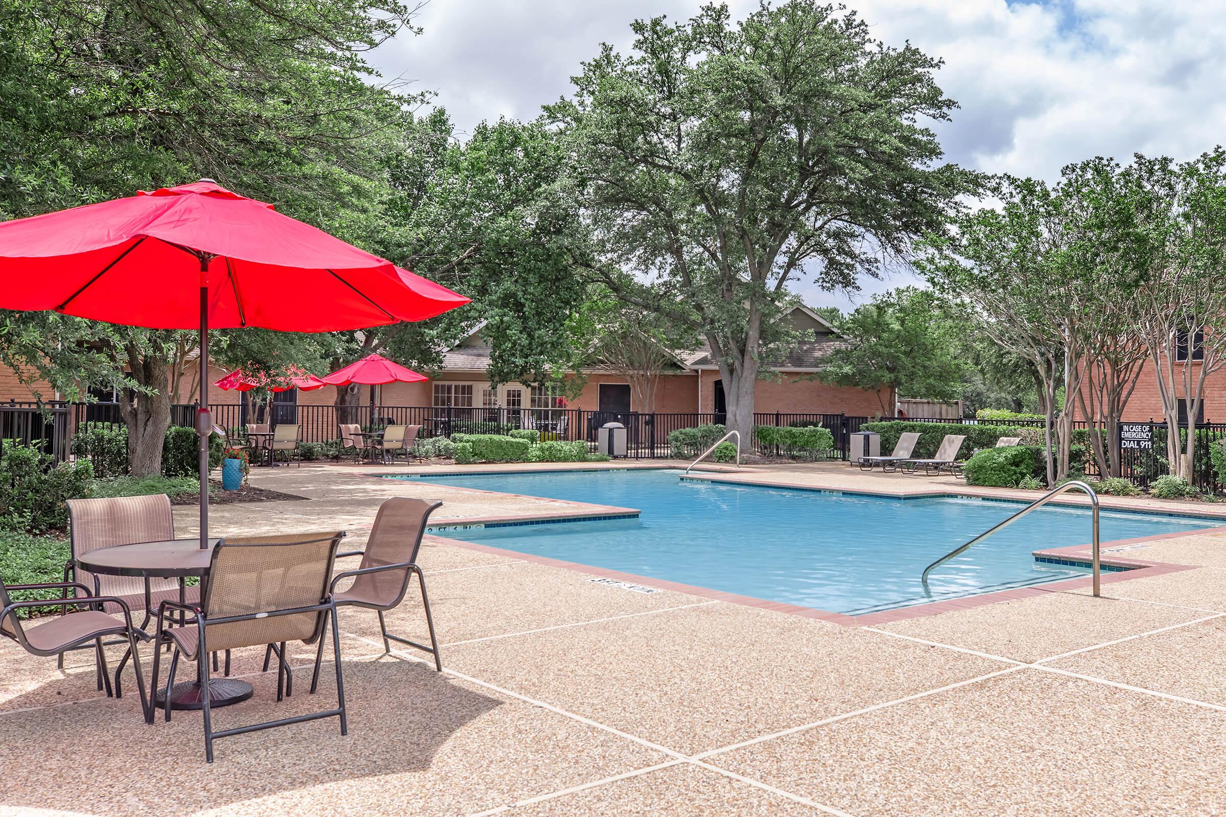 A serene outdoor swimming pool area featuring clear blue water, surrounded by lush greenery. Red umbrellas provide shade over tables and chairs, while lounge chairs are arranged along the poolside. The backdrop includes a brick building and a cloudy sky, creating a relaxing atmosphere.