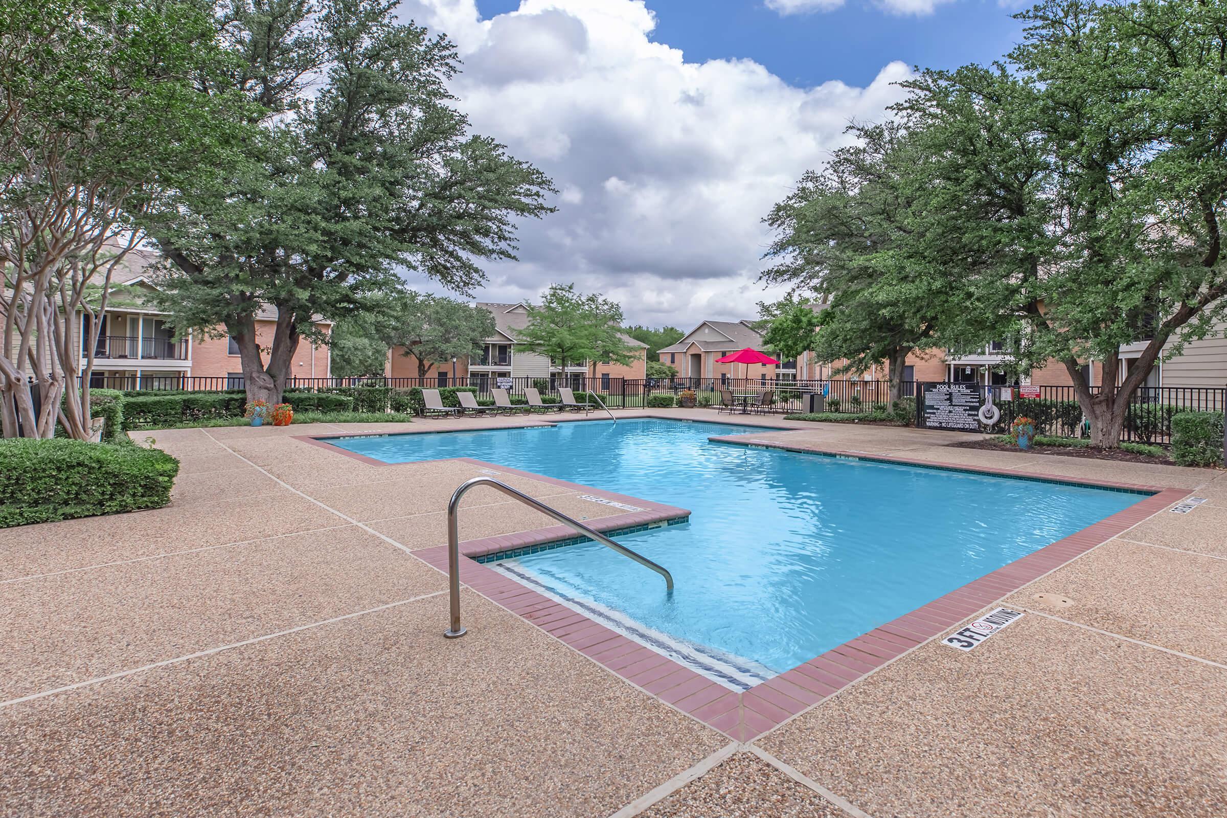 A serene swimming pool surrounded by lush greenery and residential buildings. The pool features a diving board and lounge chairs under shade. Bright blue sky with fluffy white clouds adds to the inviting atmosphere, making it a perfect spot for relaxation and leisure.