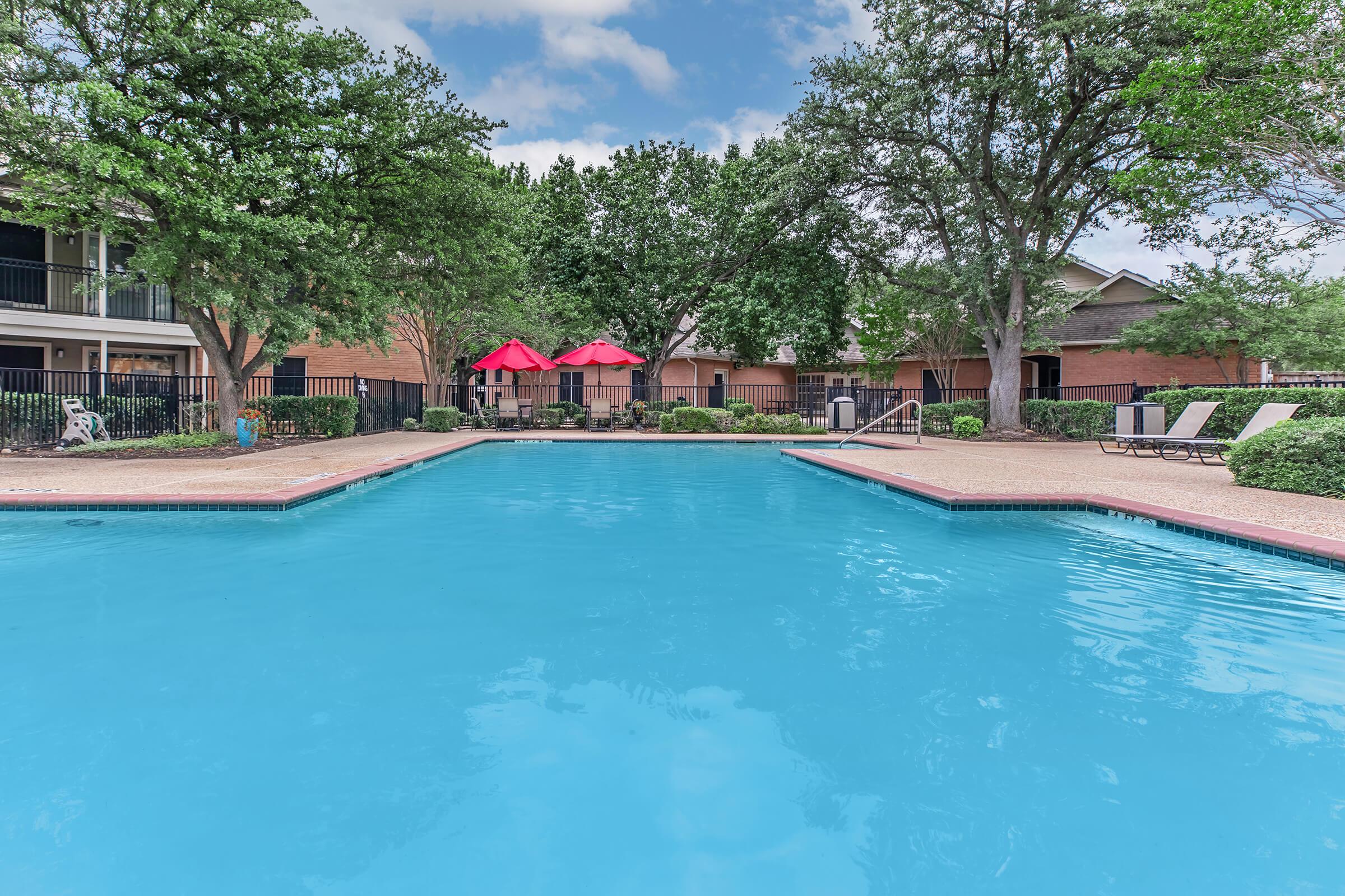 A clear, inviting swimming pool surrounded by lush greenery and trees. There are two red umbrellas and lounge chairs near the pool. The pool area is part of an apartment complex, featuring multiple buildings in the background. The sky is partly cloudy, enhancing the serene outdoor atmosphere.