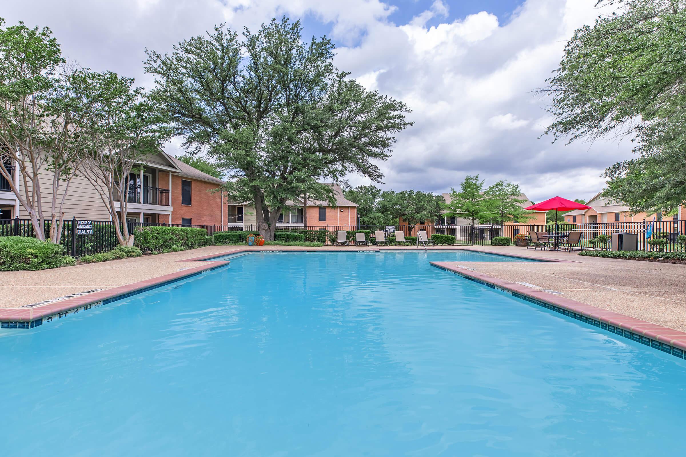 A clear blue swimming pool surrounded by well-maintained landscaping, with lounge chairs and tables nearby. Trees and residential buildings frame the scene under a partly cloudy sky. Umbrellas provide shaded areas around the pool.