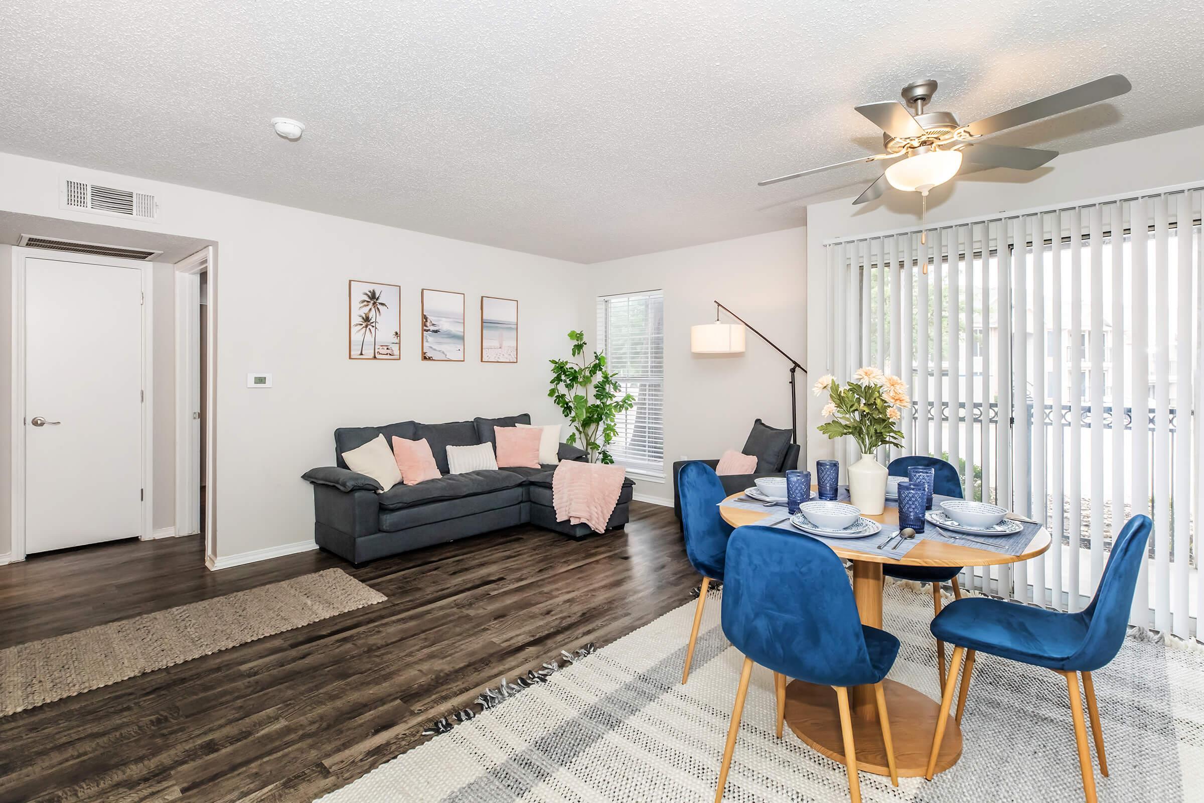 A cozy living room with a gray sofa and decorative pillows, matched with a round dining table set for four with blue chairs. There are framed beach-themed prints on the wall, a potted plant in the corner, and a bright window with vertical blinds allowing natural light.