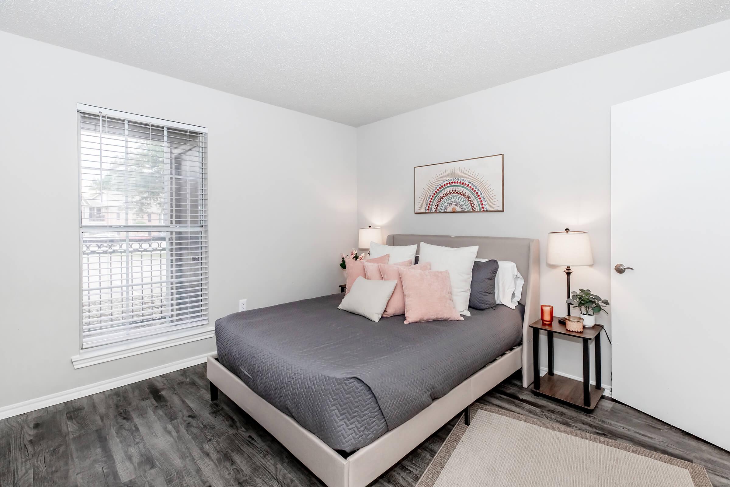 A cozy bedroom featuring a gray bed with decorative pillows, a small bedside table with candles, and a framed rainbow artwork on the wall. Natural light comes through a window with blinds, illuminating the room's neutral-colored walls and wooden floor.