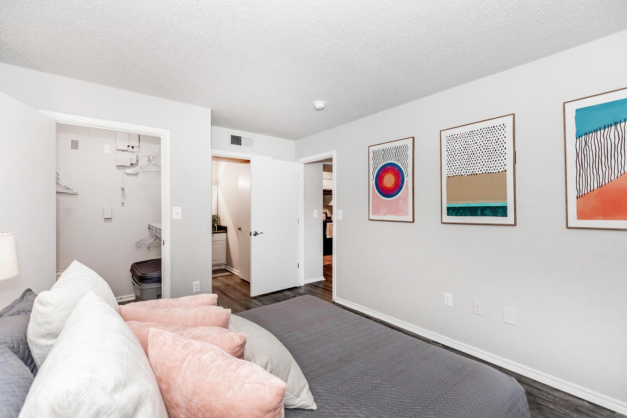 A cozy bedroom featuring a bed with decorative pillows, light-colored walls, and framed artwork on the wall. The doorway leads to another room, and a closet is visible on the left. The space has a modern and inviting atmosphere with soft lighting.