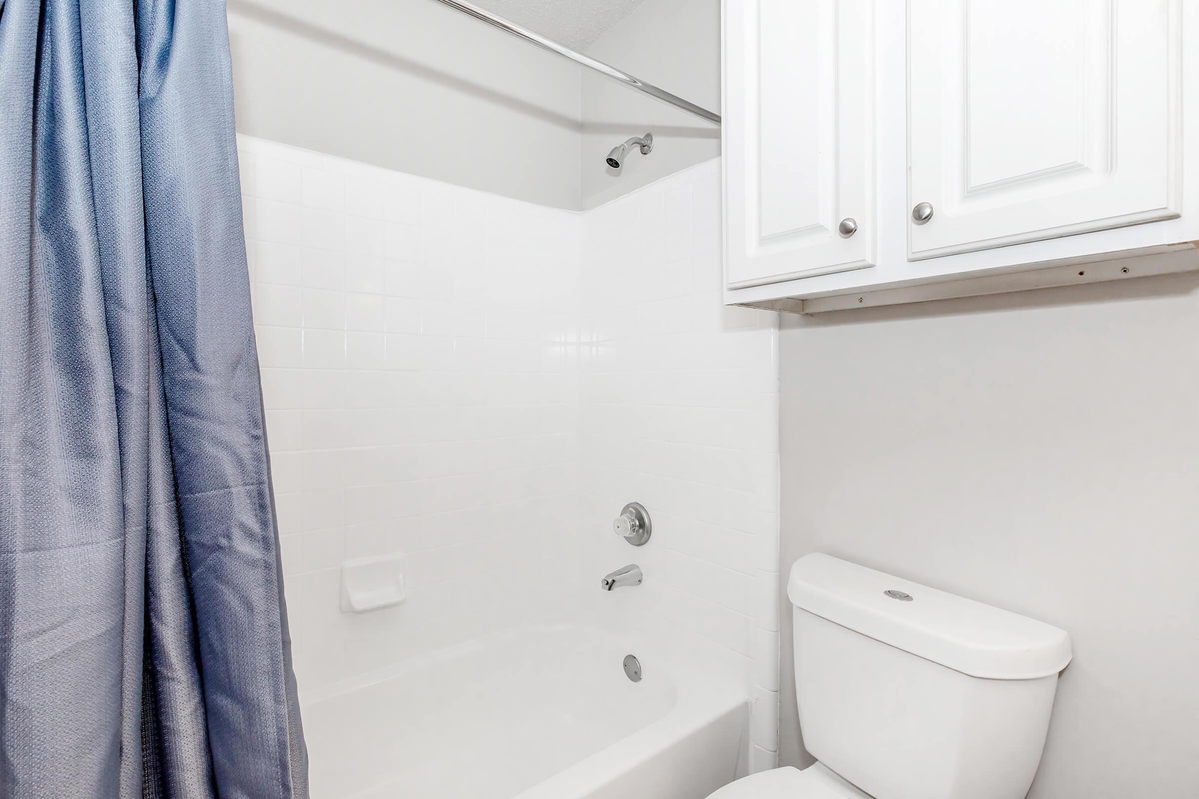 A clean and minimalistic bathroom featuring a white bathtub with a shower curtain in blue, a chrome faucet, and a matching white toilet. Above the tub, there is a glass shower rod. The walls are painted in a light color, and there's a cabinet above the toilet for storage.