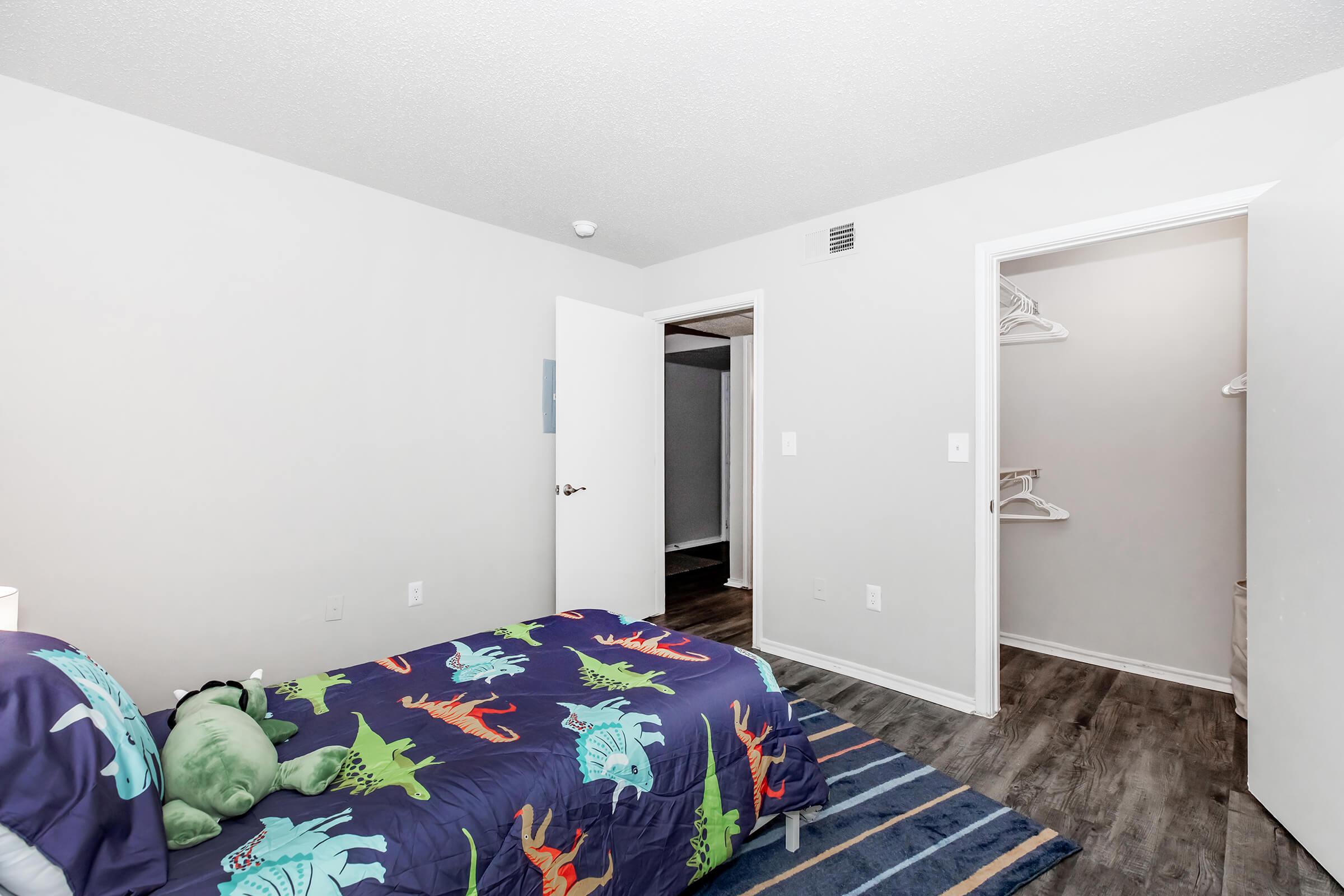 A children's bedroom featuring a colorful dinosaur-themed bedspread. The room has light gray walls and dark wooden flooring, with a closet and an open door leading to another space. A green plush toy is placed on the bed, adding to the playful atmosphere.