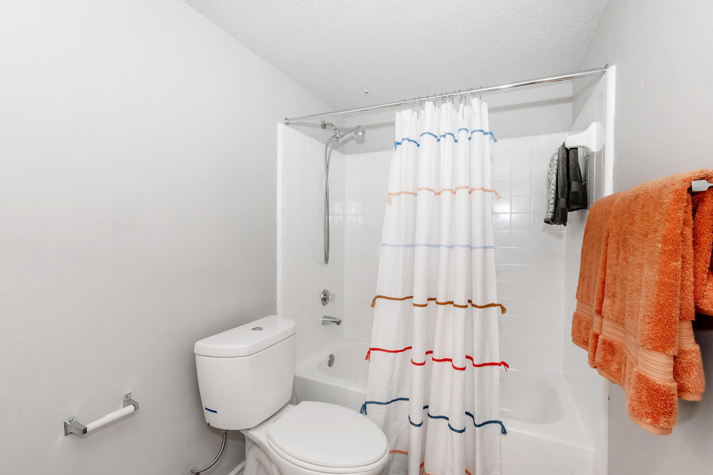 A clean bathroom featuring a bathtub with a shower, a white shower curtain with colorful stripes, a toilet, and two orange towels hanging on a rack. The walls are light gray, and the flooring is not visible.