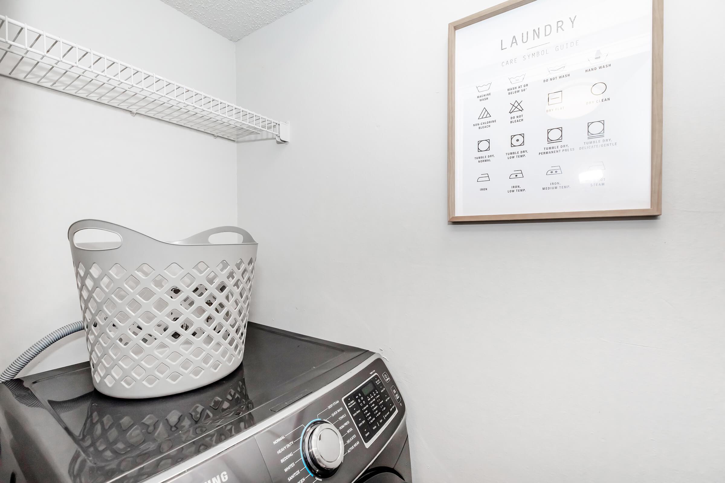 A laundry room featuring a top-loading washing machine with a control panel, a white plastic laundry basket, a wire shelf, and a framed print on the wall providing laundry care symbols and instructions. The room has neutral-colored walls and good lighting, creating a clean and organized appearance.