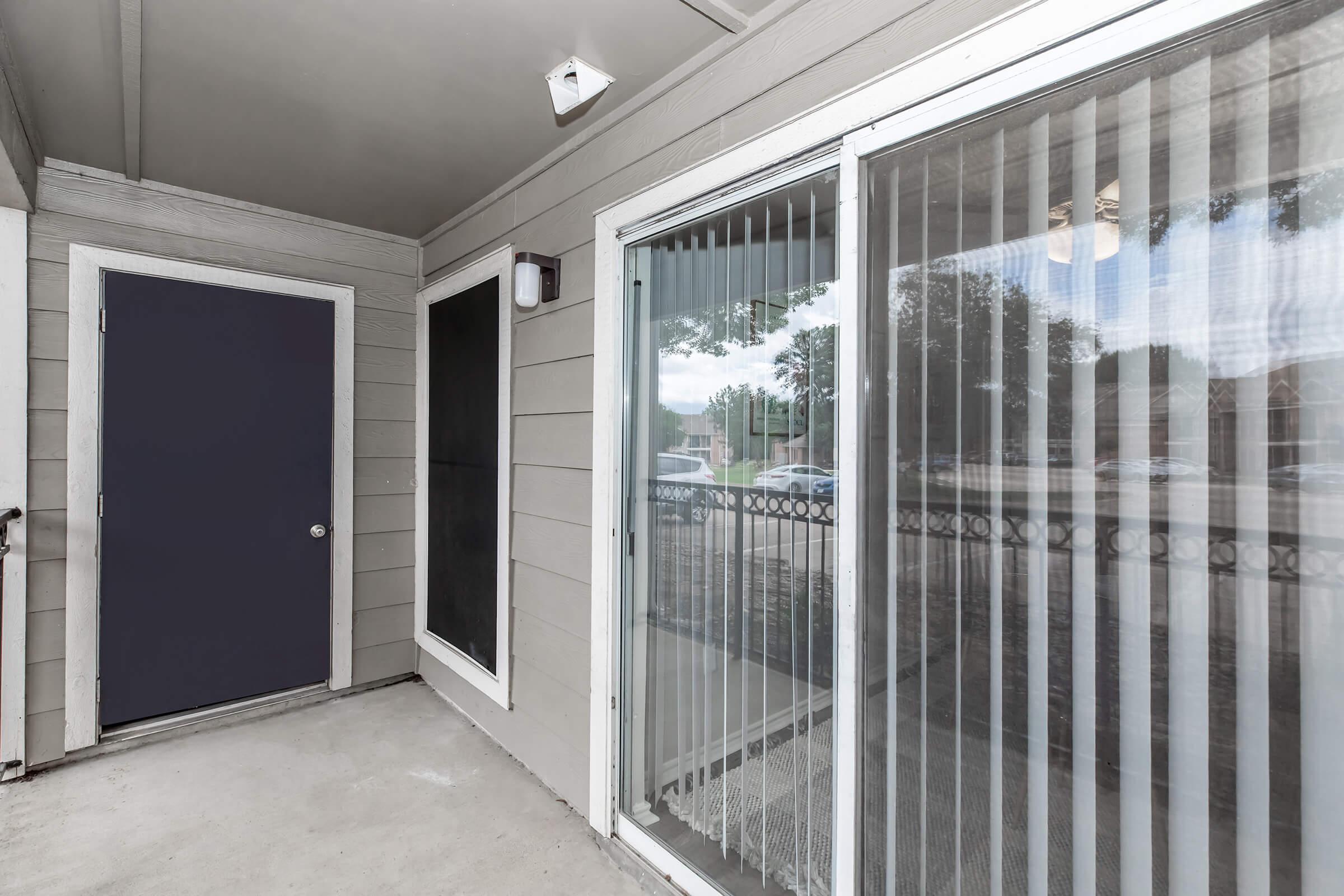 A view of a covered porch area featuring a dark blue door, a window with vertical blinds, and light gray siding. The porch is partially enclosed, and thereโs a railing visible. Natural light fills the space, creating a welcoming atmosphere.