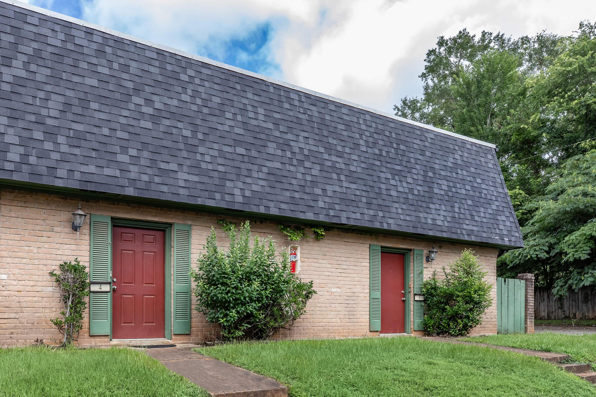 Two side-by-side entrances of a single-story, brick building with a steep, dark shingle roof. Each entrance has a red door, bordered by green shutters. Lush grass and small bushes are in front of the building, with trees visible in the background under a partly cloudy sky.