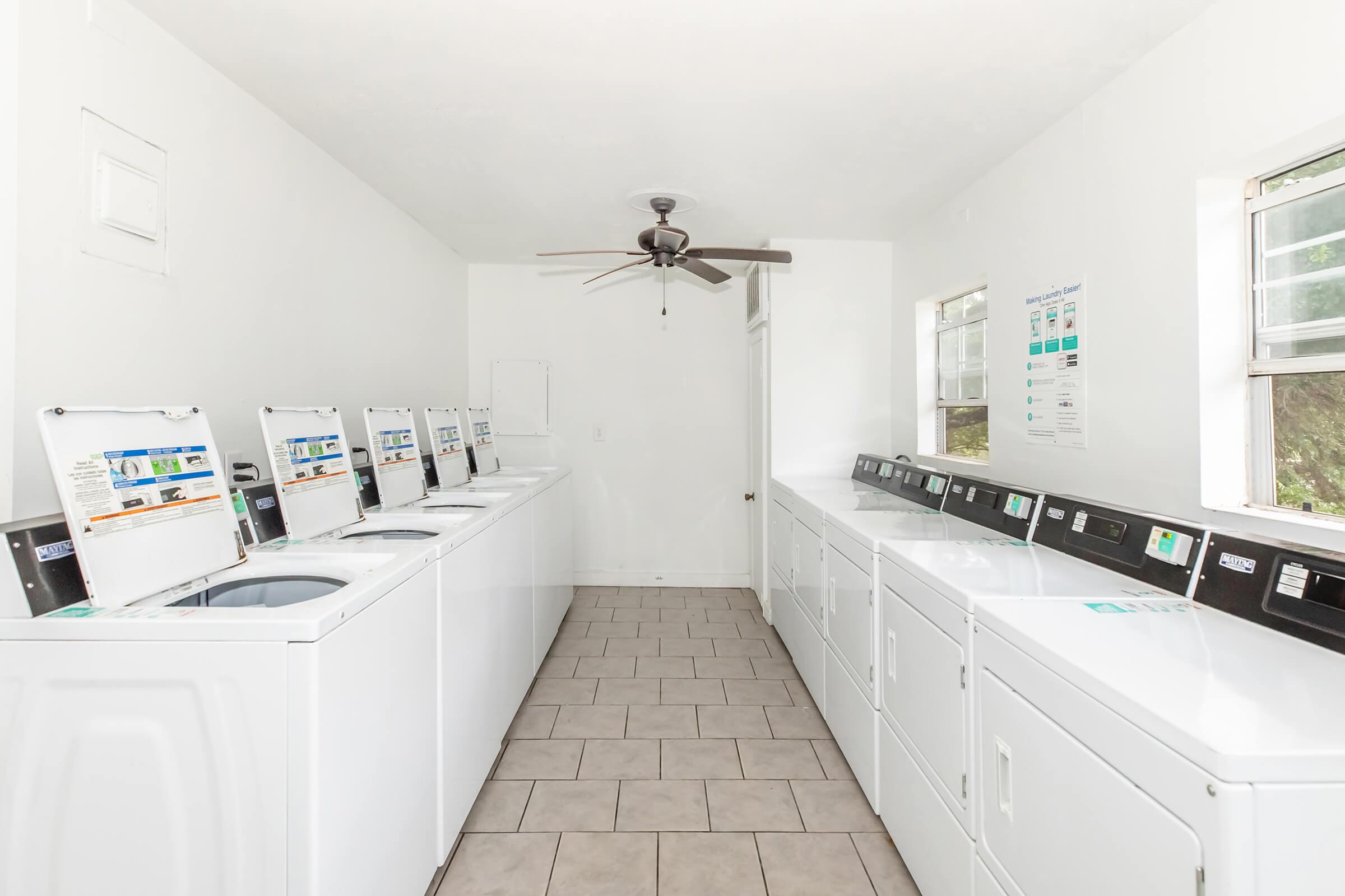 A clean, well-lit laundry room featuring several white washing machines and dryers lined along the walls. There are windows allowing natural light, a ceiling fan for ventilation, and tiled flooring, creating a functional and inviting space for laundry tasks.