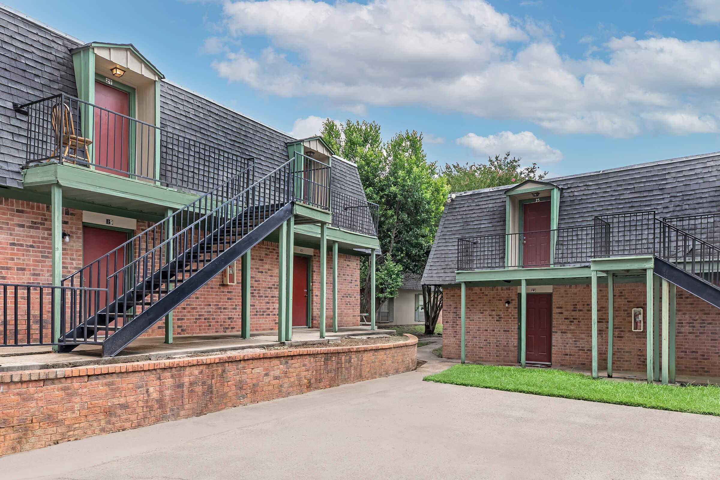 Two buildings with brick exteriors and green trim, featuring multiple staircases leading to second-floor entrances. The scene includes well-maintained grass and a clear blue sky with clouds. The architecture reflects a classic multi-unit layout, creating a welcoming residential environment.