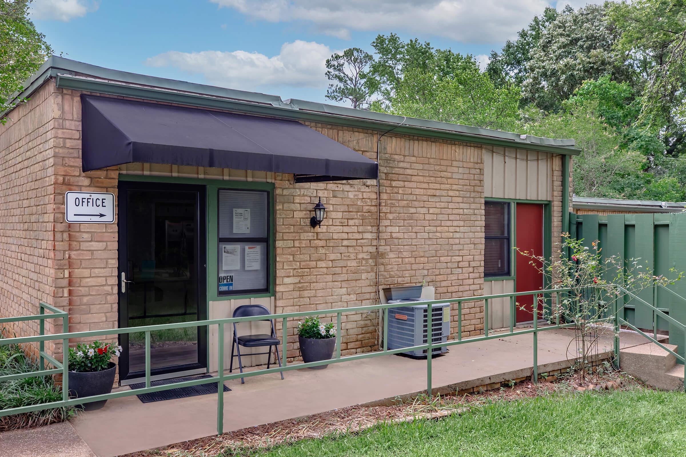 Single-story office building with a brick façade, black awning, and a sign labeled "OFFICE." Entrance features a small porch with potted plants and a chair. Surrounding area has green grass and trees, with another building visible to the right.