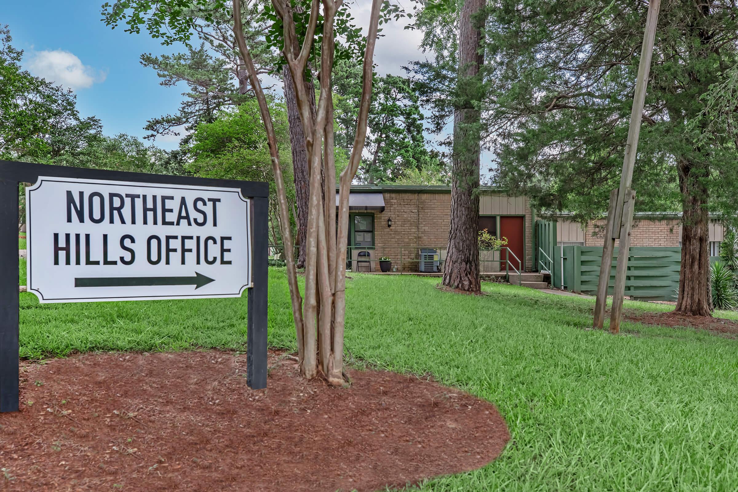 Sign for "Northeast Hills Office" with an arrow pointing left, located in a grassy area surrounded by trees. The office building is visible in the background, with a pathway leading up to it.