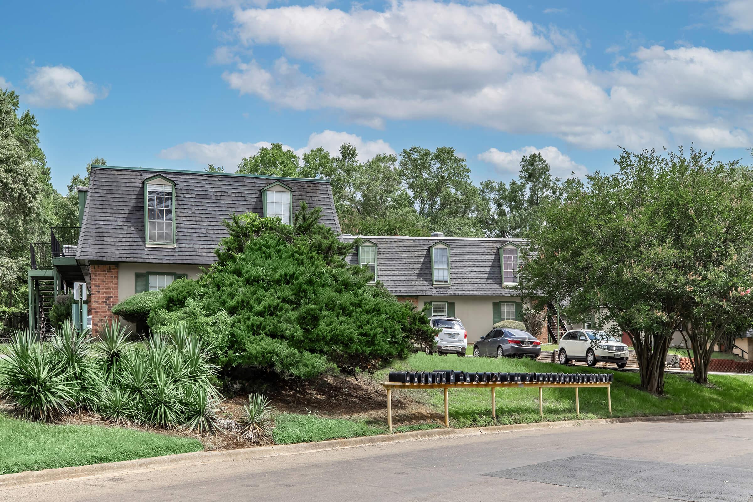 A residential neighborhood featuring a two-story house with a sloped roof and large windows. In the foreground, there are landscaped bushes and trees, while a driveway holds multiple parked cars. The sky above is partly cloudy, adding a pleasant ambiance to the scene.