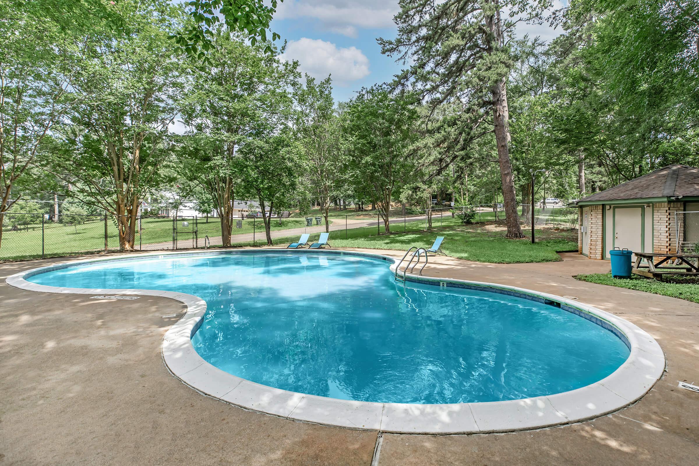 A clear blue swimming pool with a curvy shape, surrounded by green trees and grass. There are two lounge chairs near the pool, and a small building with a shed-like structure nearby. The scene is bright and inviting, suggesting a peaceful outdoor recreational area.
