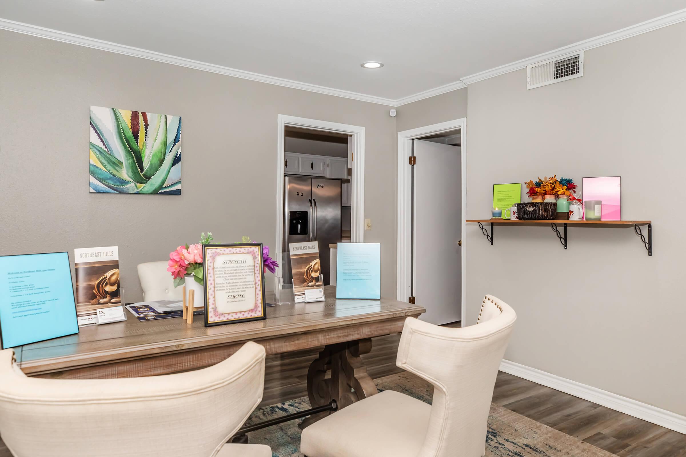 A modern office space featuring a wooden table surrounded by two cream-colored chairs. On the table, there are various brochures and a vase with flowers. The walls are painted light gray, with a colorful piece of art depicting an aloe plant and a small shelf displaying decorative items and flyers.