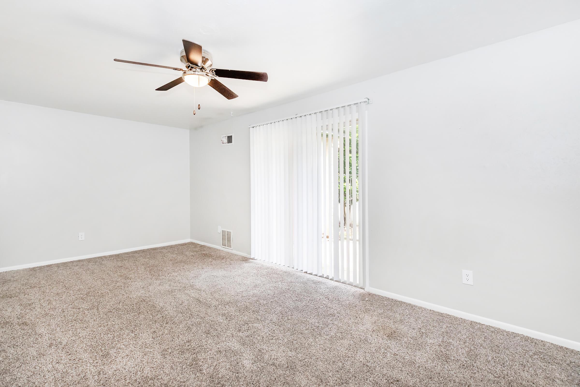 A spacious living room featuring light gray walls, soft beige carpet, and a ceiling fan. Large sliding glass doors with vertical blinds allow natural light to enter, leading to an outdoor space. The room has an open, airy feel with minimal furnishings.