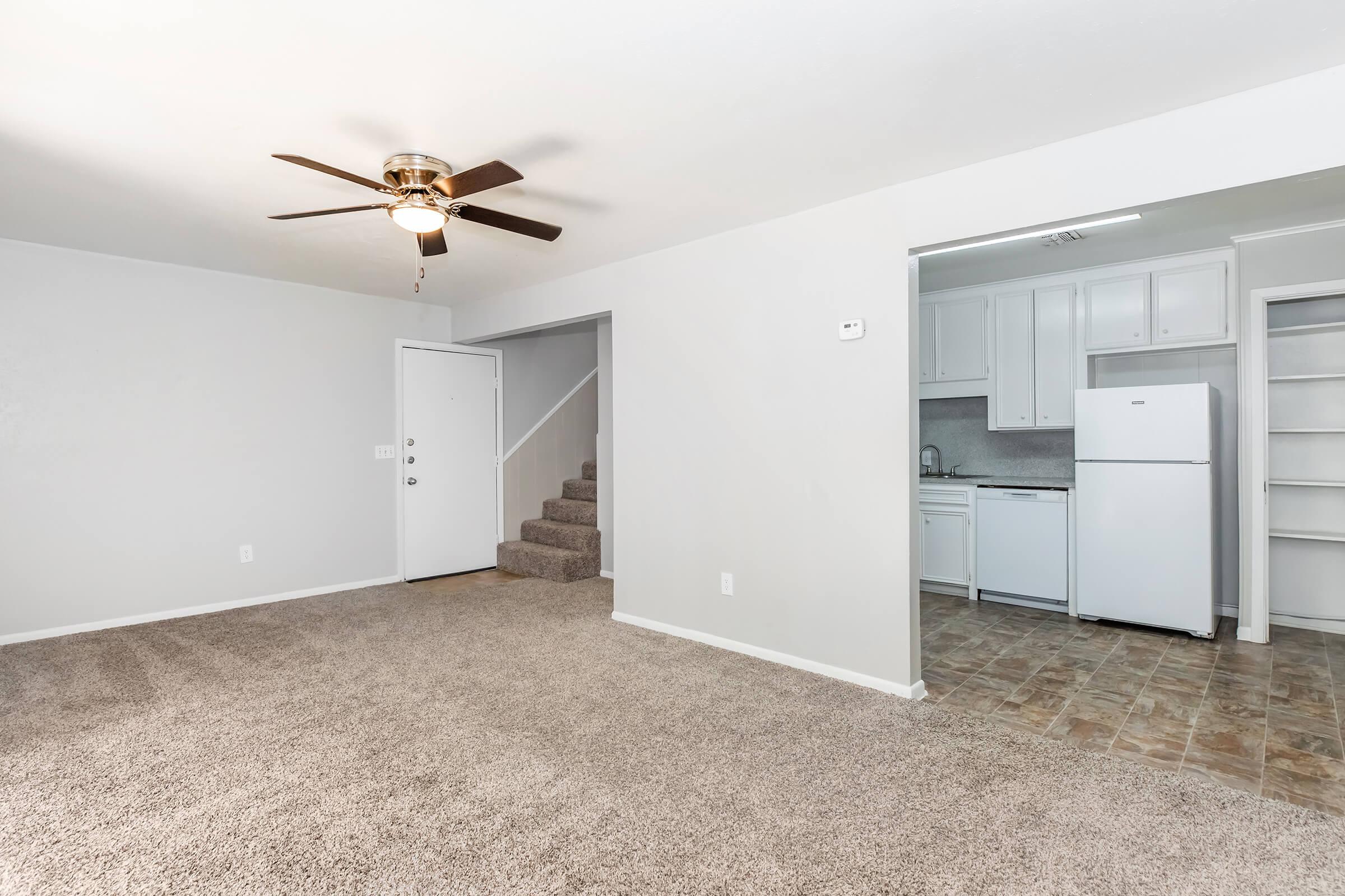 A spacious living area with neutral-colored walls and plush carpet, featuring a ceiling fan. To the right, a doorway leads into a kitchen area, visible with white cabinetry and a refrigerator. A staircase is located in the background, enhancing the open feel of the space.