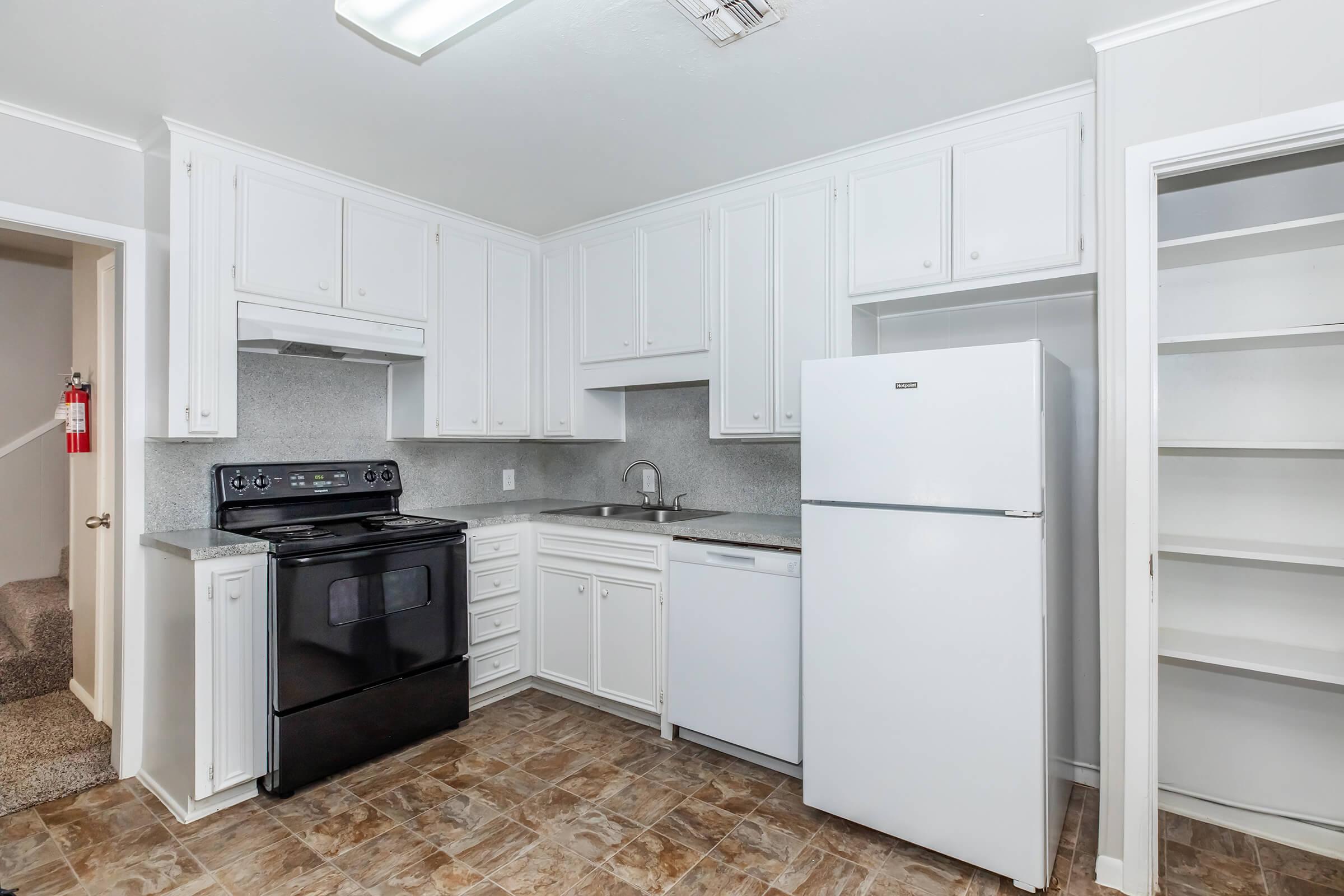 A clean and modern kitchen featuring white cabinetry, a black stove and oven, a white refrigerator, and a dishwasher. The space has light-colored walls, vinyl flooring, and a pantry with open shelving. A door leads to an adjoining room, and there is overhead lighting in the ceiling.