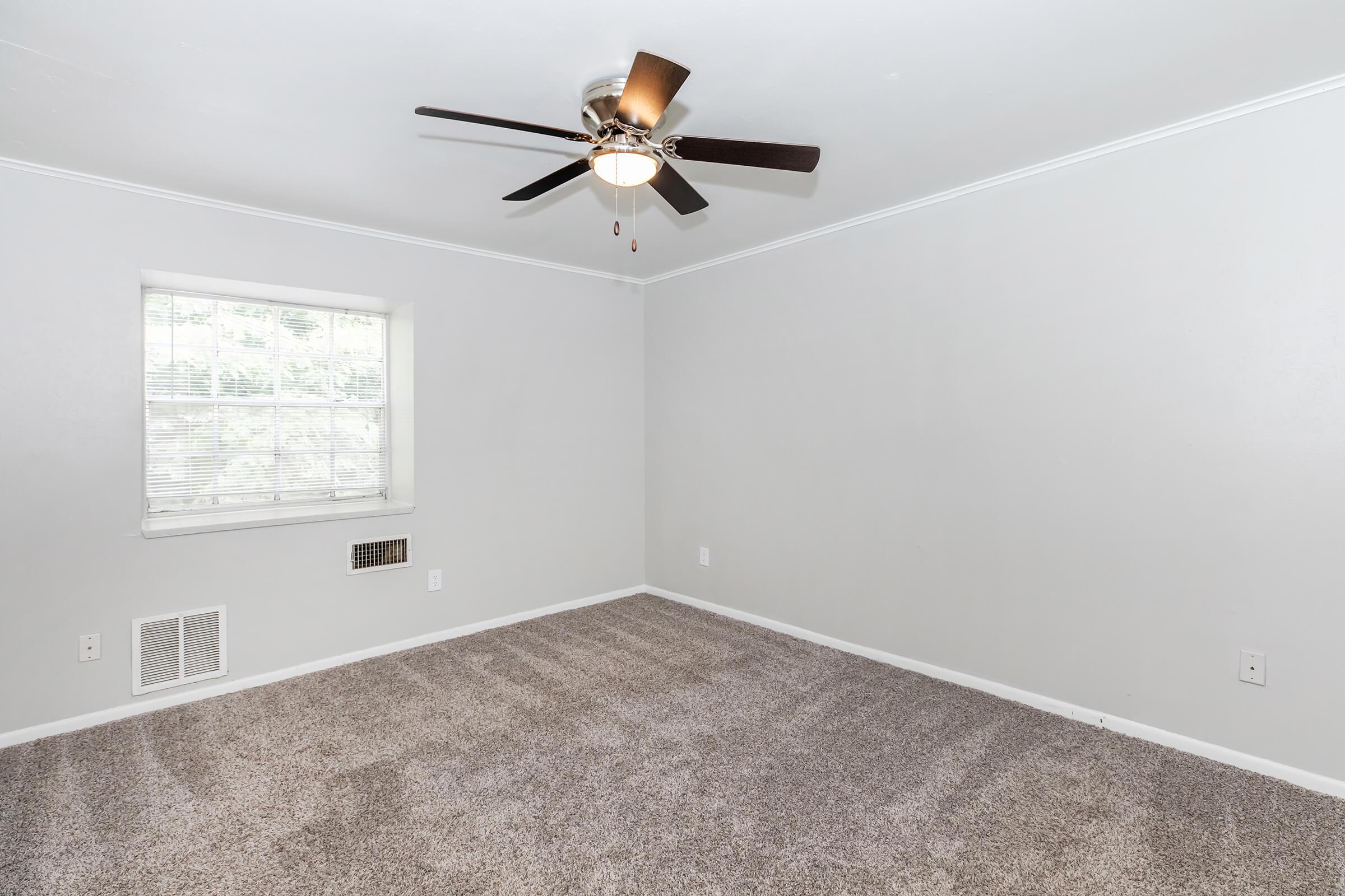 A vacant room with light gray walls and plush carpet. It features a ceiling fan with five blades, a window with blinds that lets in natural light, and an air vent on the wall. The space is minimalistic, offering a clean, open area suitable for various purposes.