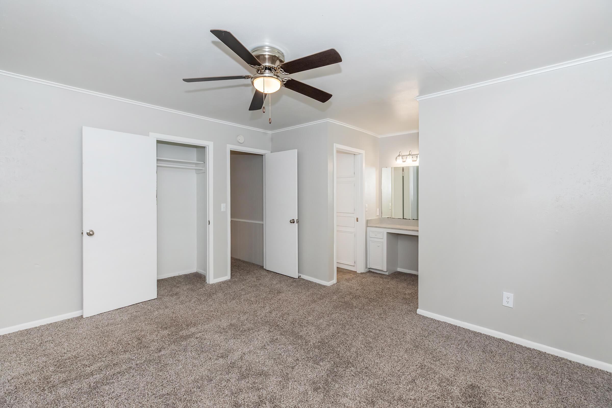 A spacious room featuring light gray walls, plush carpeting, and a ceiling fan. Two white double doors lead to a closet, while a bathroom area with a vanity and mirror is visible on the right. The overall atmosphere is bright and inviting, ideal for a bedroom or personal space.