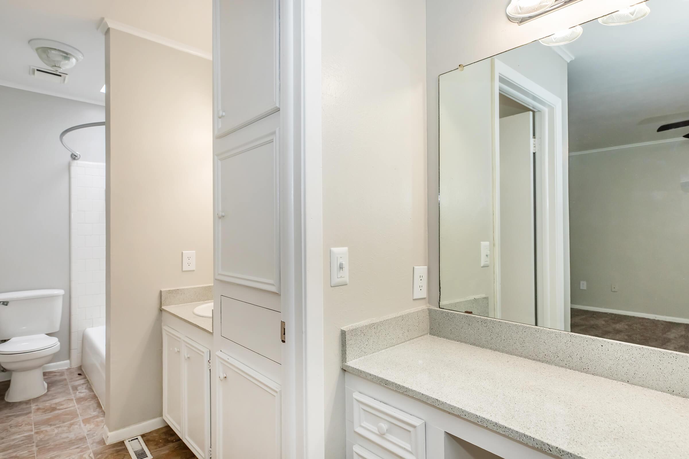 Bright and minimalist bathroom space featuring a white vanity with a gray countertop, a large mirror, and beige walls. A bathtub with a shower curtain is visible on the left, while a toilet is positioned nearby. Soft natural light enhances the clean and modern look of the room.