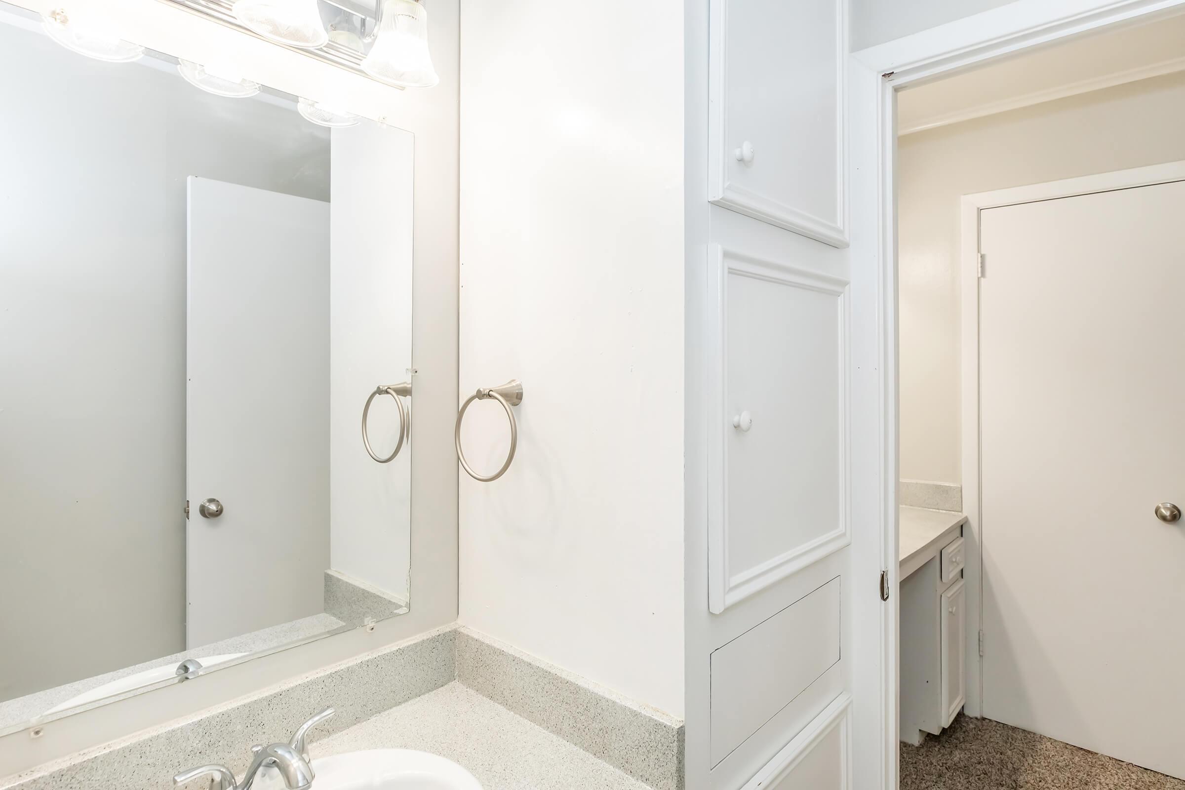 A clean and simple bathroom featuring a countertop sink with a mirror above, a towel ring on the wall, and a doorway leading to another room. The walls are painted white, and the floor appears to have a textured surface. A bright, well-lit atmosphere is created by overhead lighting.