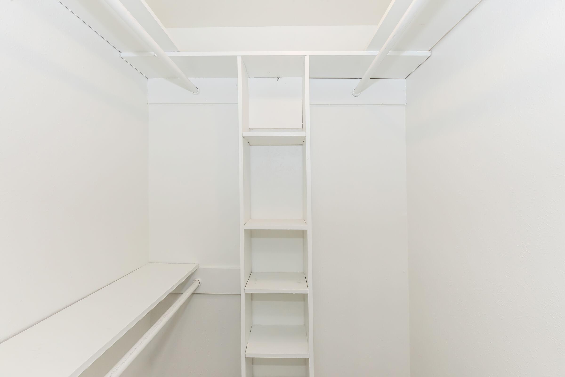 Empty white closet with two horizontal rods for hanging clothes and a vertical section of shelves on one side. The walls and shelving are also white, creating a clean and minimalist appearance.