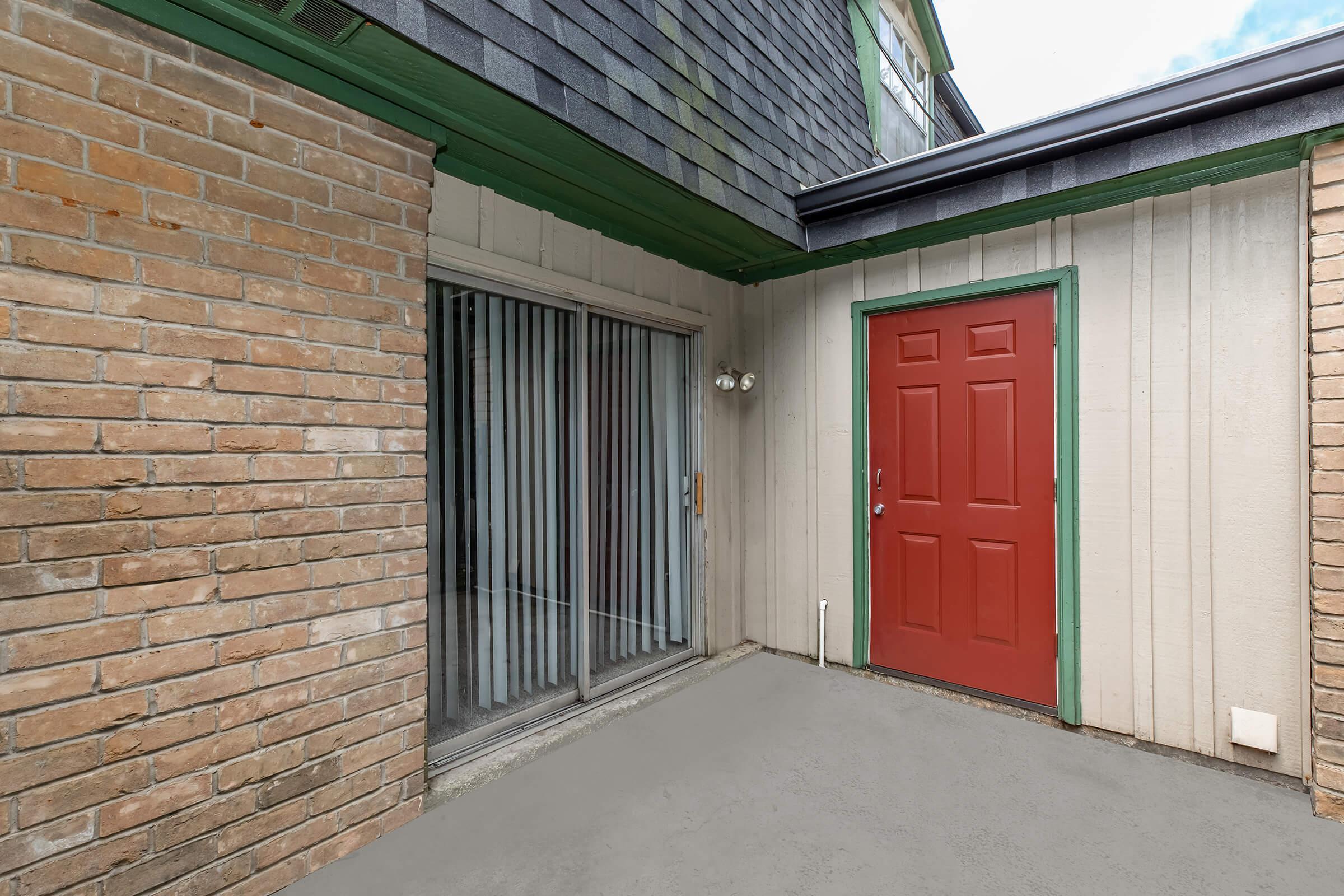 A partially covered entrance featuring a red door and adjacent sliding glass door with vertical blinds. The walls are a mix of brick and textured siding, with a concrete floor. A green trim outlines the roof, and there are cloudy skies visible above.