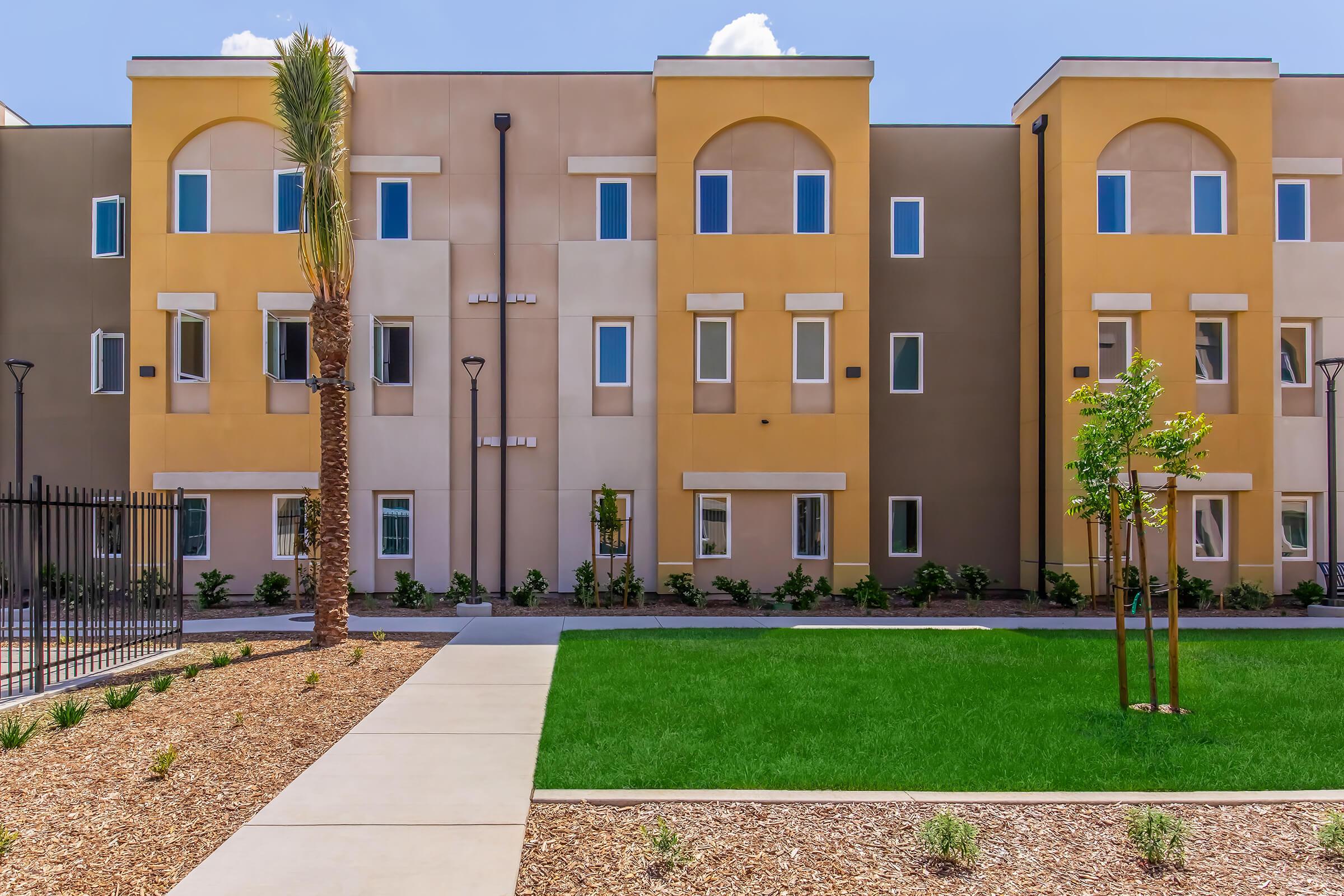 Modern residential building with multiple stories featuring yellow and beige facades, large windows, and palm trees. A landscaped area with green grass and decorative gravel paths is in the foreground, enhancing the inviting atmosphere of the property. Bright blue sky with a few clouds above.