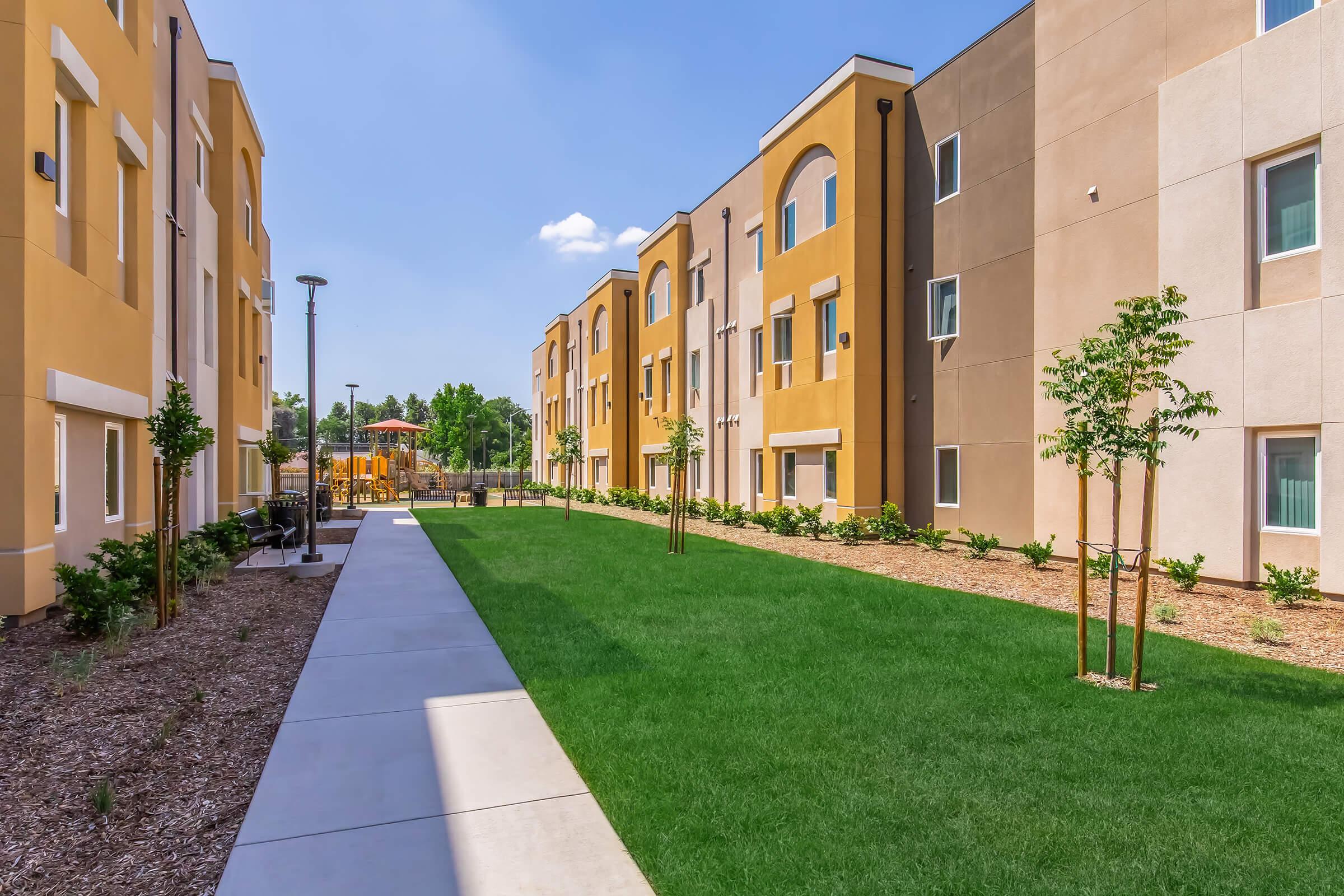 A well-maintained outdoor area featuring a pathway lined with green grass and young trees, flanked by modern apartment buildings. In the background, there is a playground visible, with clear blue skies enhancing the bright and inviting atmosphere of the space.
