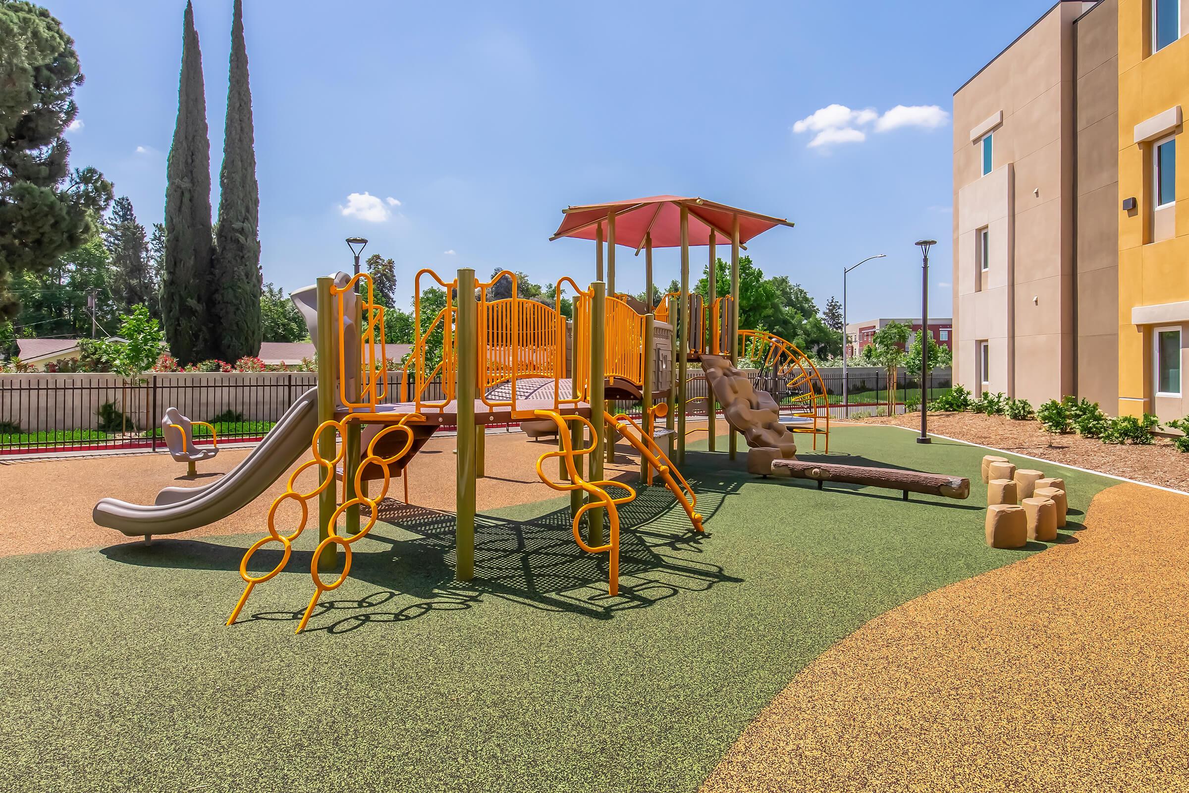 A colorful playground featuring a yellow climbing structure with slides, swings, and a rock wall. The area is surrounded by green grass and trees, with a clear blue sky in the background and a modern building to one side. Ideal for children to play and explore.