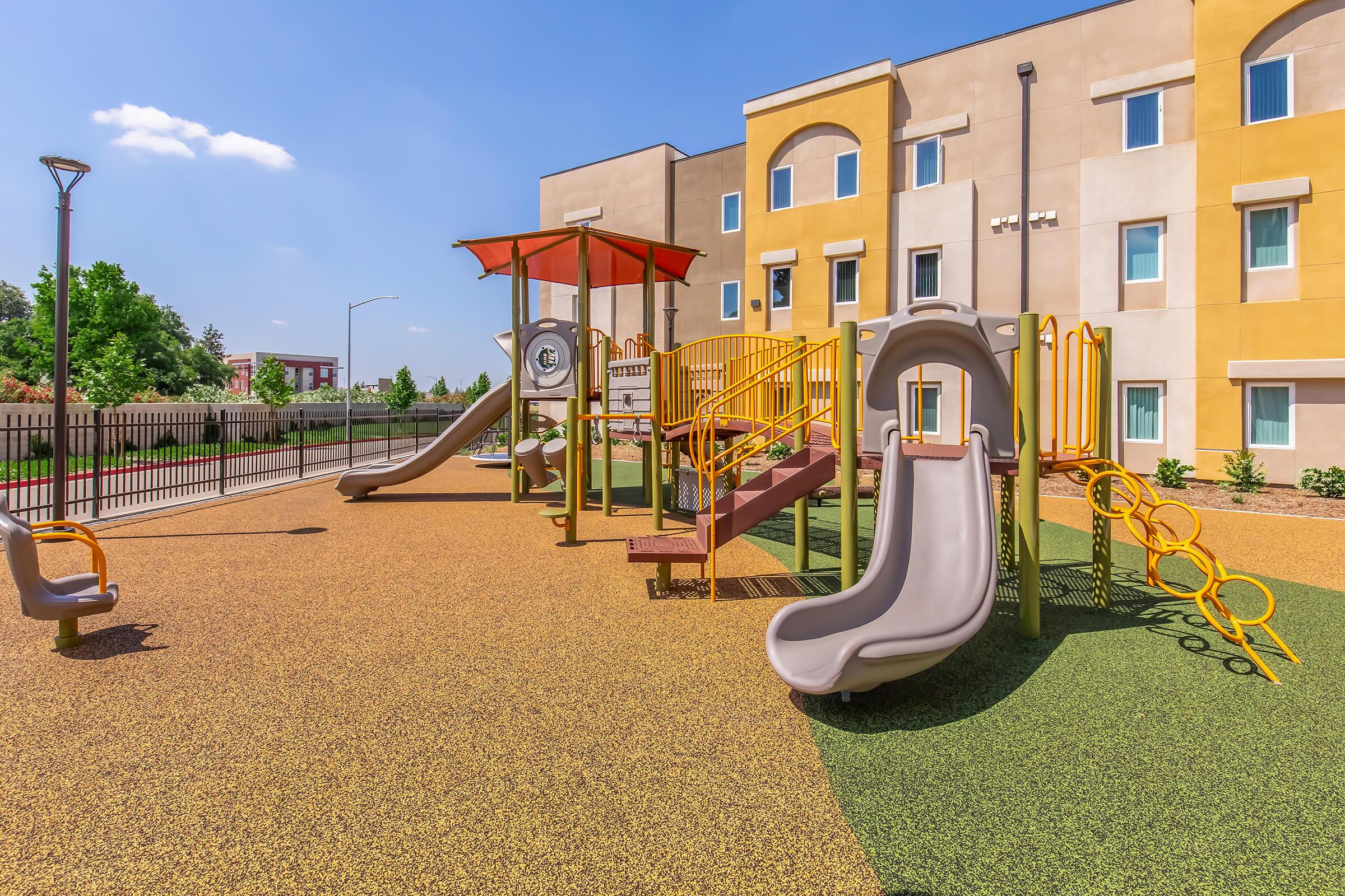 A colorful playground featuring slides, climbing structures, and swings, set on a sunny day. The playground is surrounded by a fence and located next to a modern building with yellow and beige walls. Soft ground cover provides a safe play area.