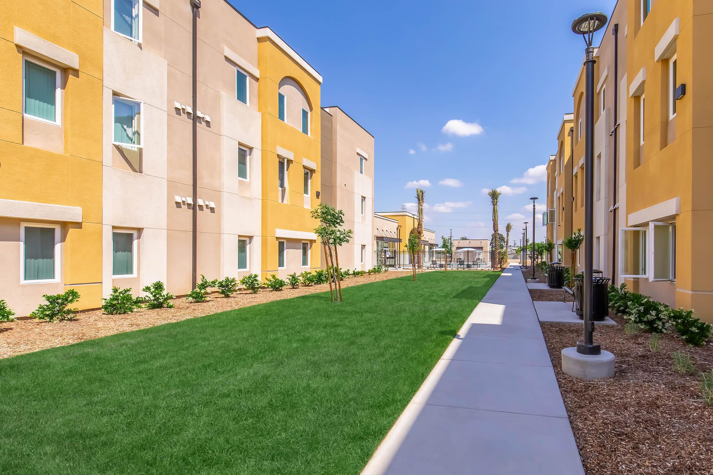 A well-maintained green pathway running between modern residential buildings. The buildings feature a mix of yellow and beige exteriors, with large windows and small trees lining the walkway. Clear blue skies and scattered clouds provide a bright atmosphere. Ideal for community living or outdoor activities.