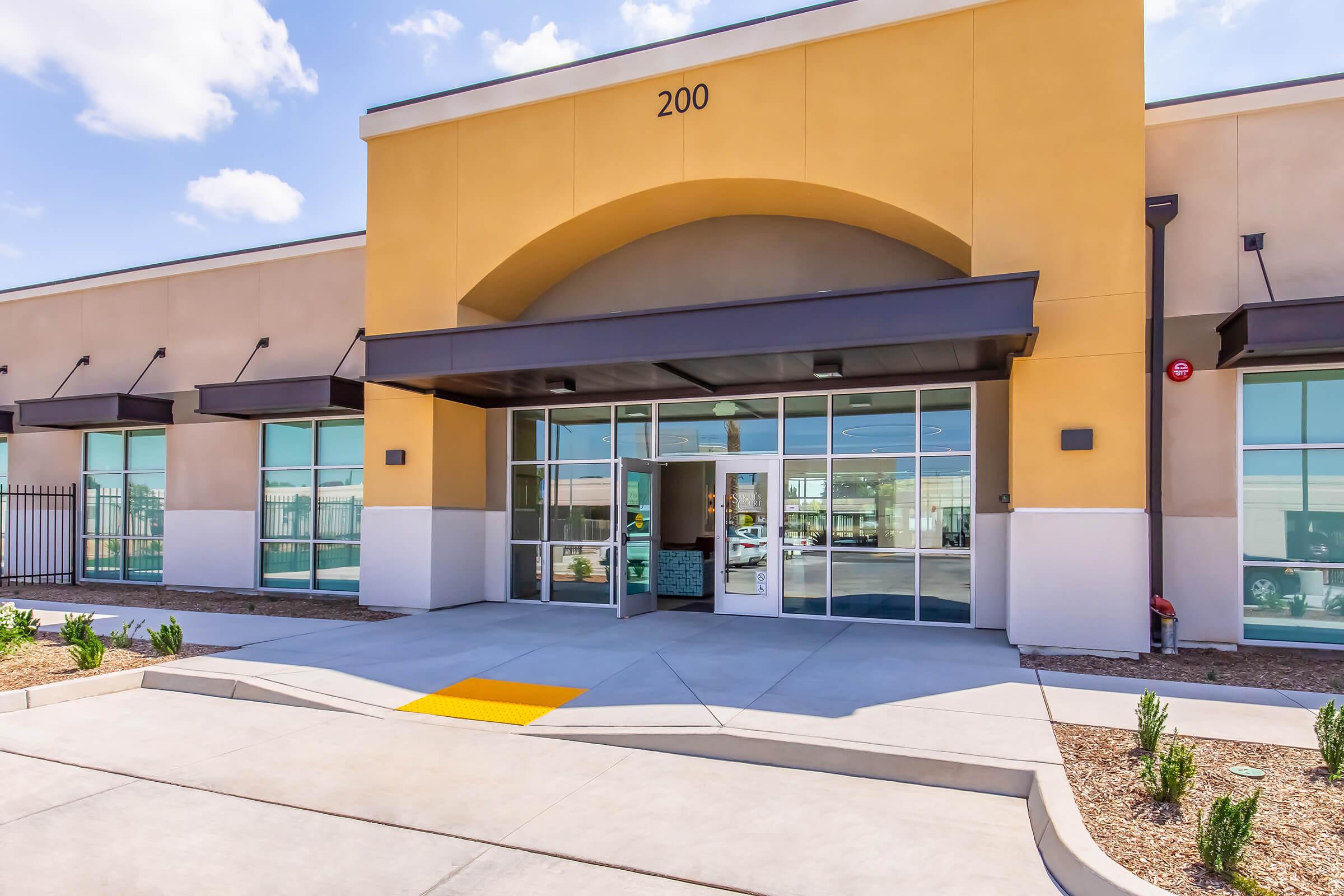 Modern building entrance featuring large glass doors, a high arched canopy, and a bright exterior. The pathway is accessible with a yellow ramp, surrounded by landscaped areas with small plants. Clear blue skies are visible above, enhancing the inviting atmosphere of the location.