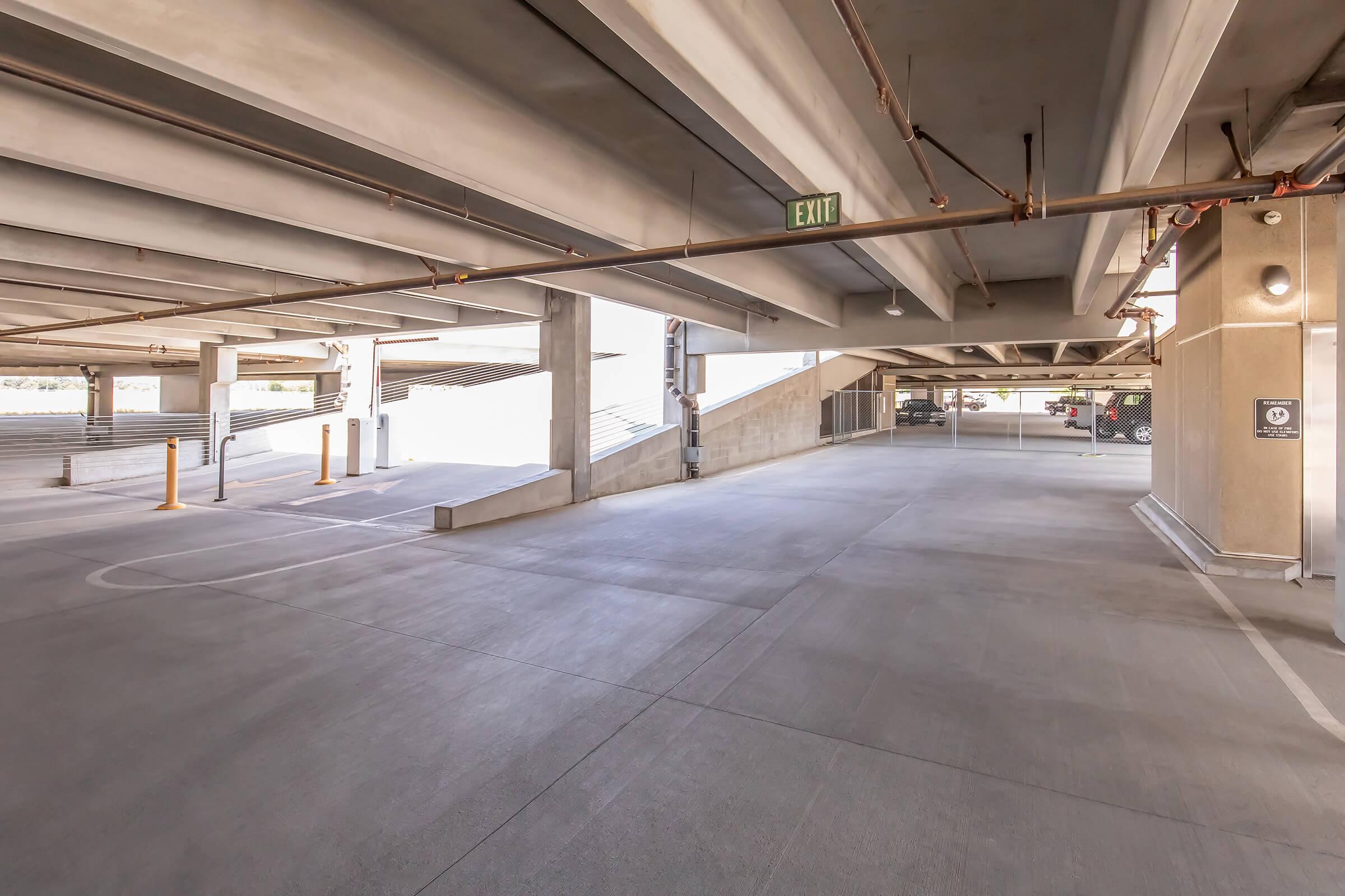 A wide view of an empty parking garage, featuring concrete walls and ceilings with exposed pipes. The lighting is bright, and there are exit signs visible. A smooth, clean floor leads to a ramp, while nearby pillars are marked with safety indicators. The overall atmosphere is spacious and well-maintained.