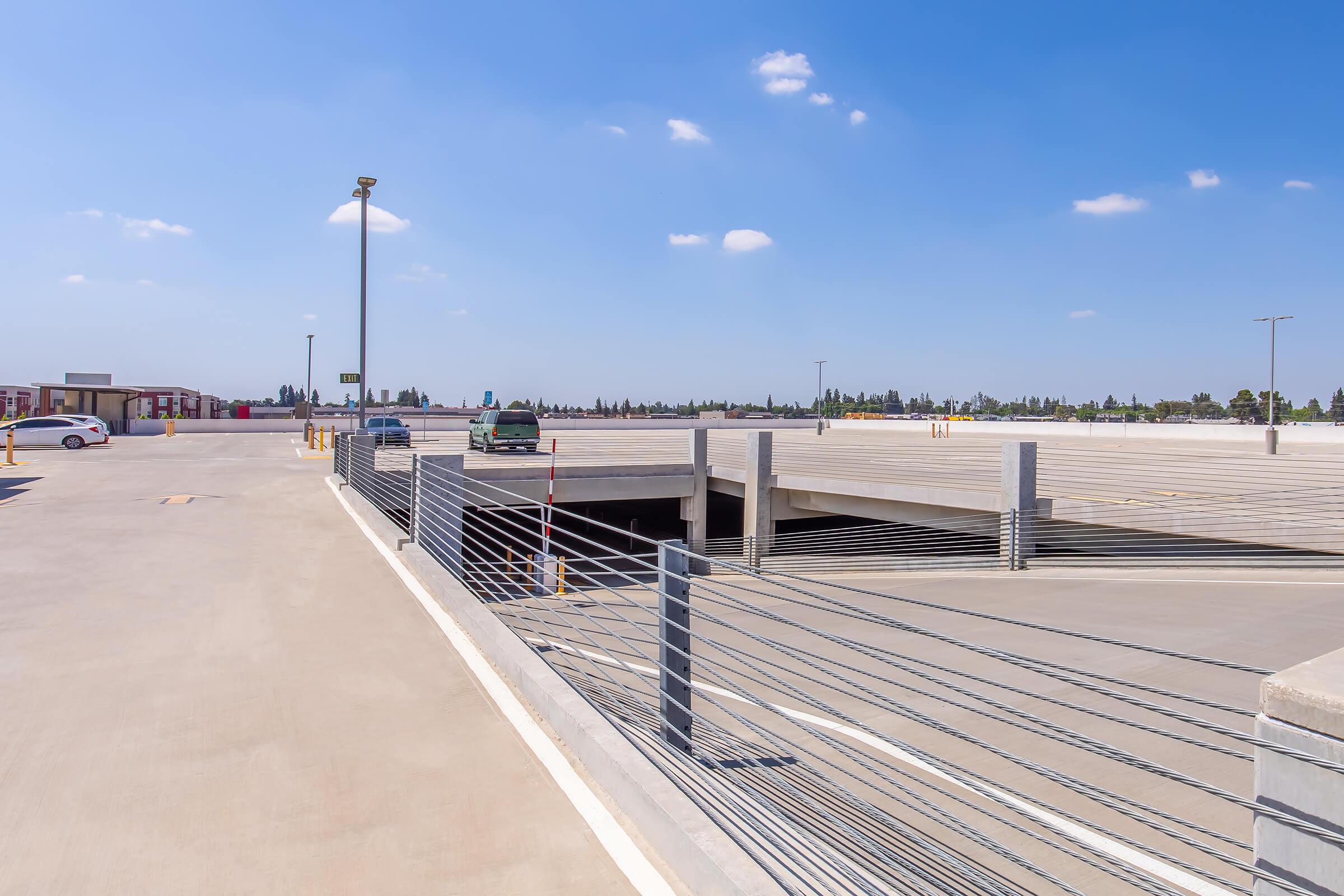 A view of a modern multi-level parking lot on a clear day. The asphalt surface is well-maintained, with vehicles parked and an entrance to an underground parking area visible. Bright blue skies and a few scattered clouds create a sunny atmosphere.
