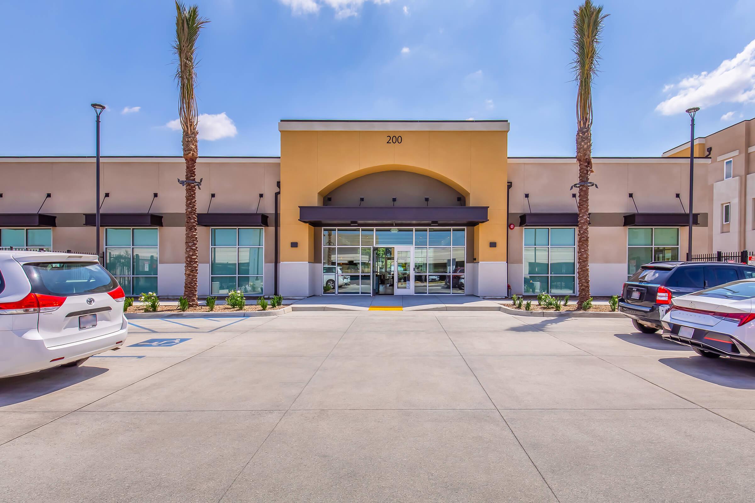 Modern commercial building facade with a central entrance, flanked by large windows. The building features an arched architectural detail above the door, and palm trees are planted on either side of the entrance. A parking lot in the foreground shows several cars parked in front. Bright blue sky with scattered clouds above.
