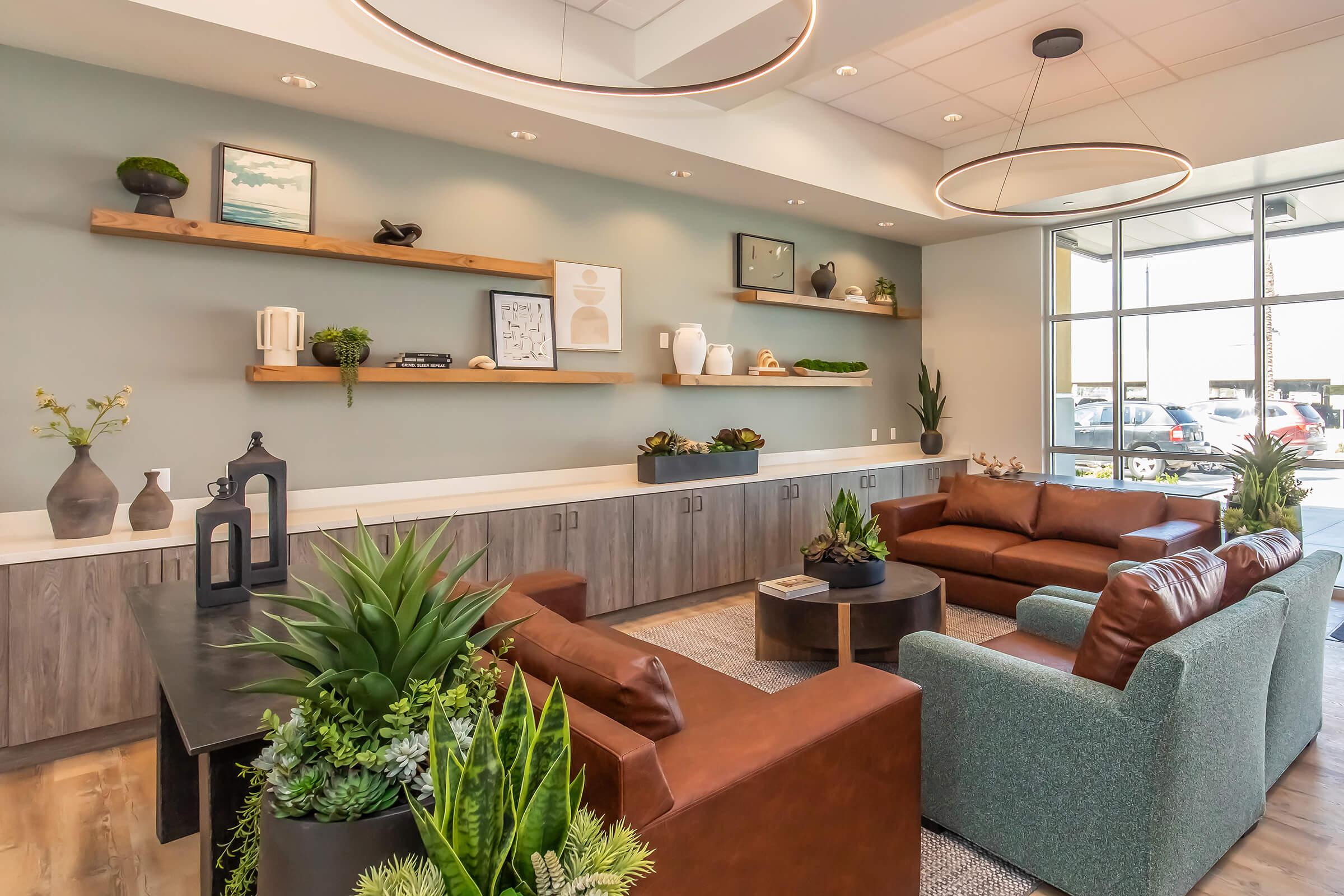 A modern lounge space featuring two leather sofas, a green upholstered chair, a coffee table, and shelves adorned with decorative items and plants. Large windows allow natural light to flood the room, creating a warm and inviting atmosphere.