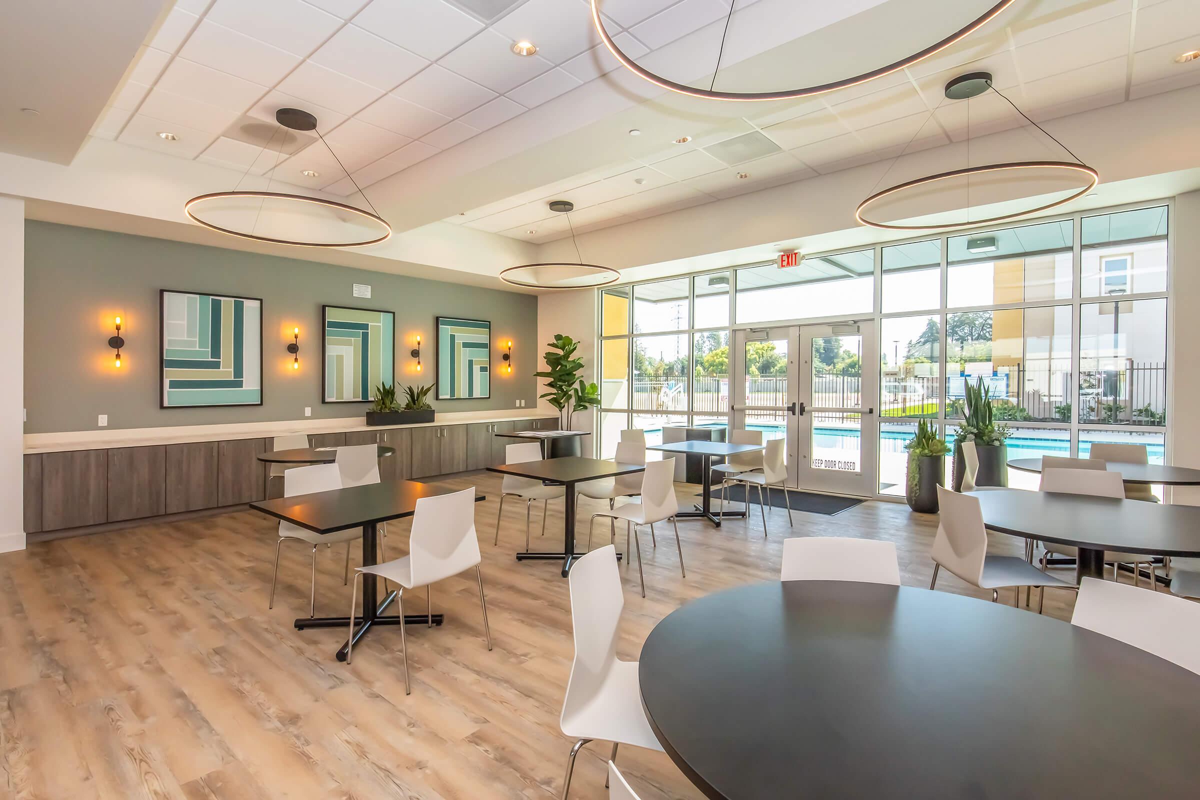 Interior of a modern communal space featuring tables and chairs, large windows allowing natural light, and decorative wall art. The area has a sleek design with pendant lighting, plants, and a view of an outdoor pool area.
