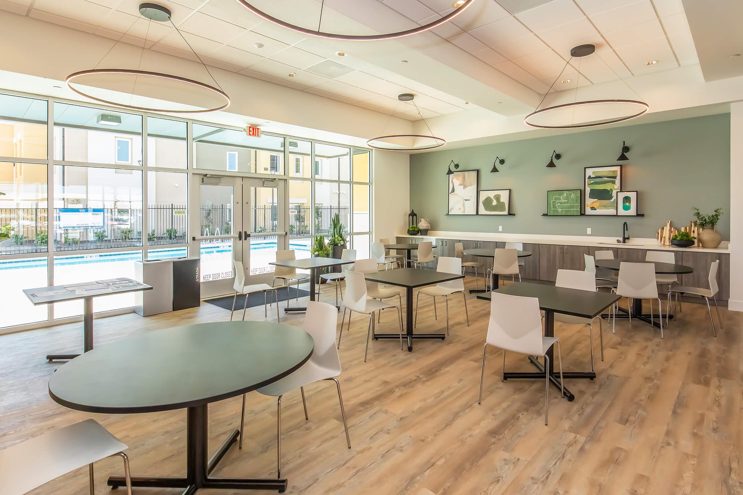 A modern dining area with multiple round tables and white chairs, featuring large windows that provide natural light and a view of an outdoor pool. The walls are painted in soft green, adorned with framed artwork. There is a sleek countertop along one side with decorative items and plants.