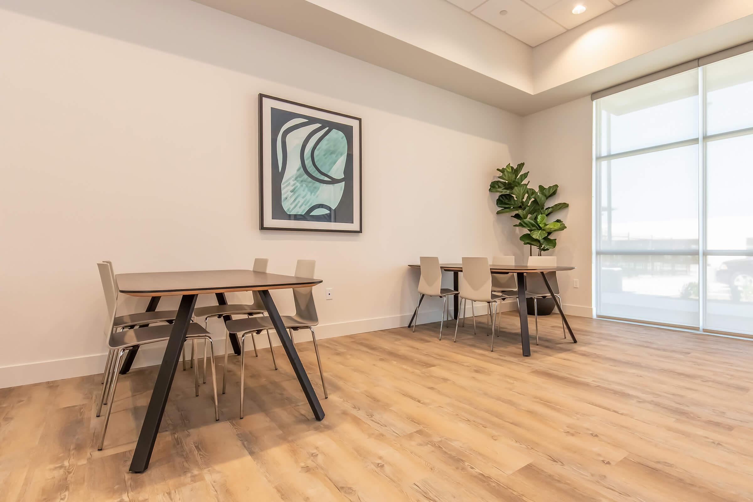 A modern, minimalistic room featuring two tables with chairs, large windows allowing natural light, and a green plant in the corner. The wall is adorned with an abstract framed artwork, complementing the light wood flooring and neutral color scheme.