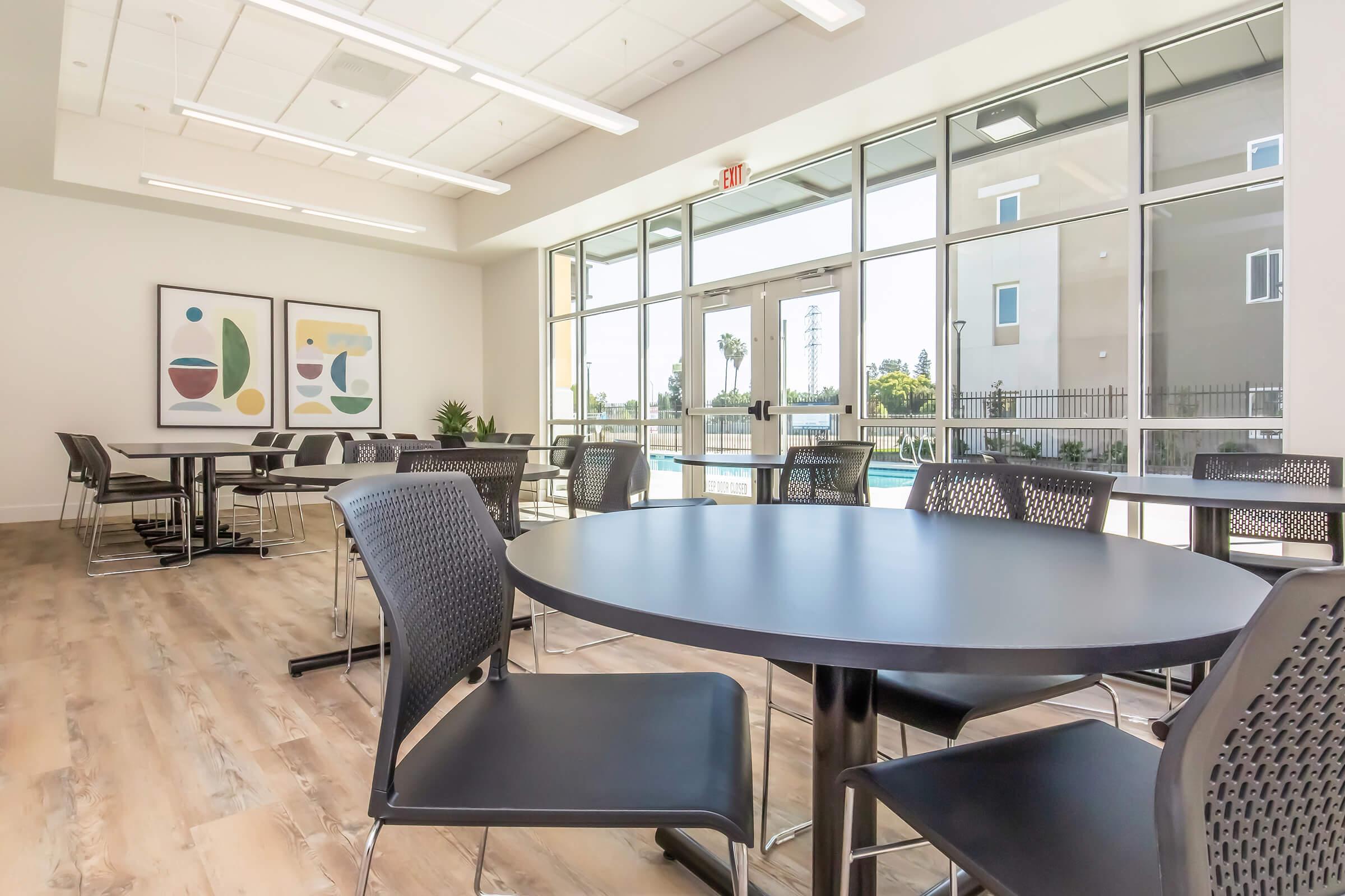 Contemporary interior of a spacious dining area featuring several round black tables surrounded by black chairs. Large windows allow natural light, revealing an outside view. Two abstract art pieces adorn the walls, and a small plant adds a touch of greenery to the space.