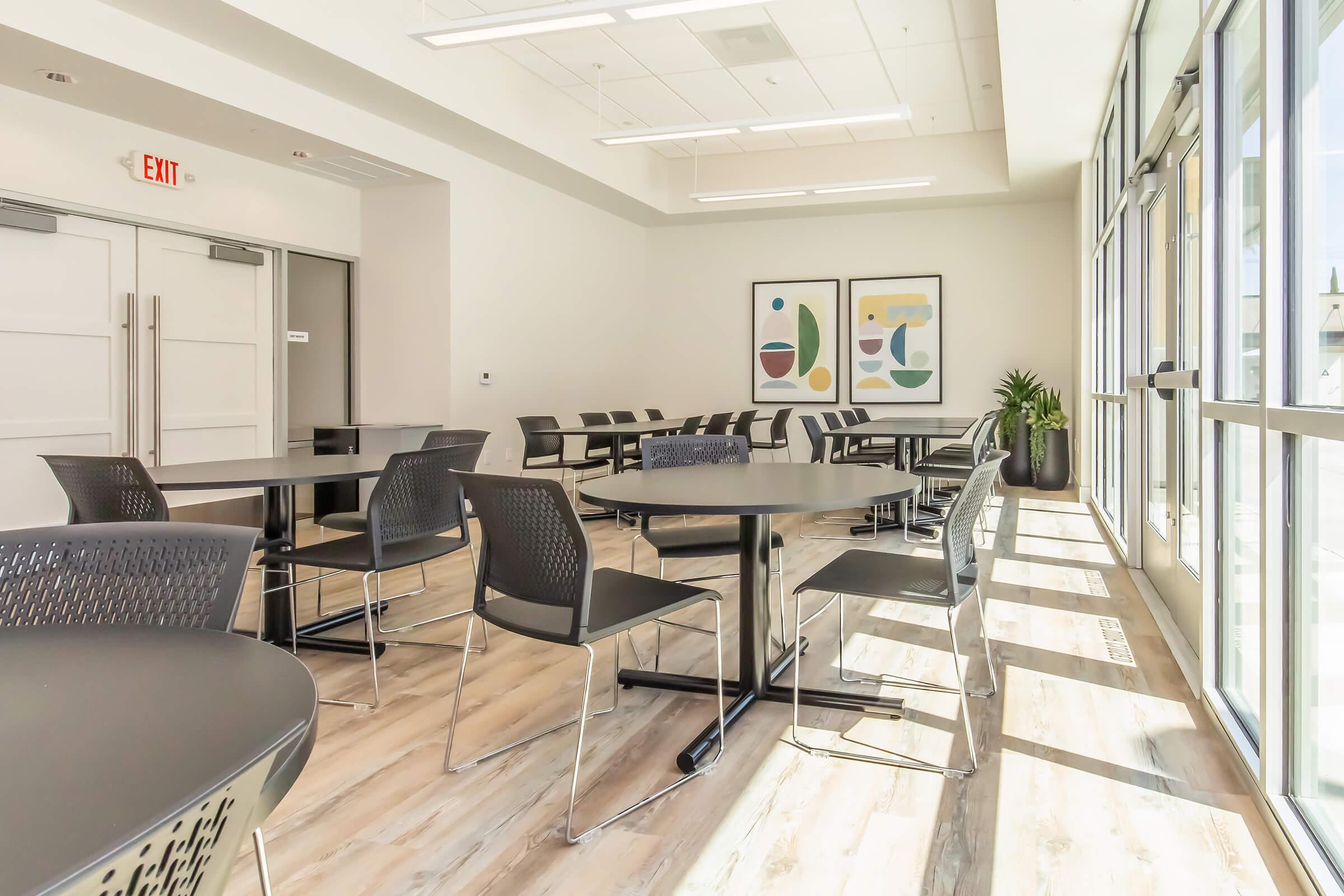 Bright, modern empty meeting room with multiple black tables and chairs arranged neatly. Large windows allow natural light to fill the space, highlighting two abstract framed artworks on the wall. A green plant is placed near the windows, adding a touch of nature to the contemporary setting.