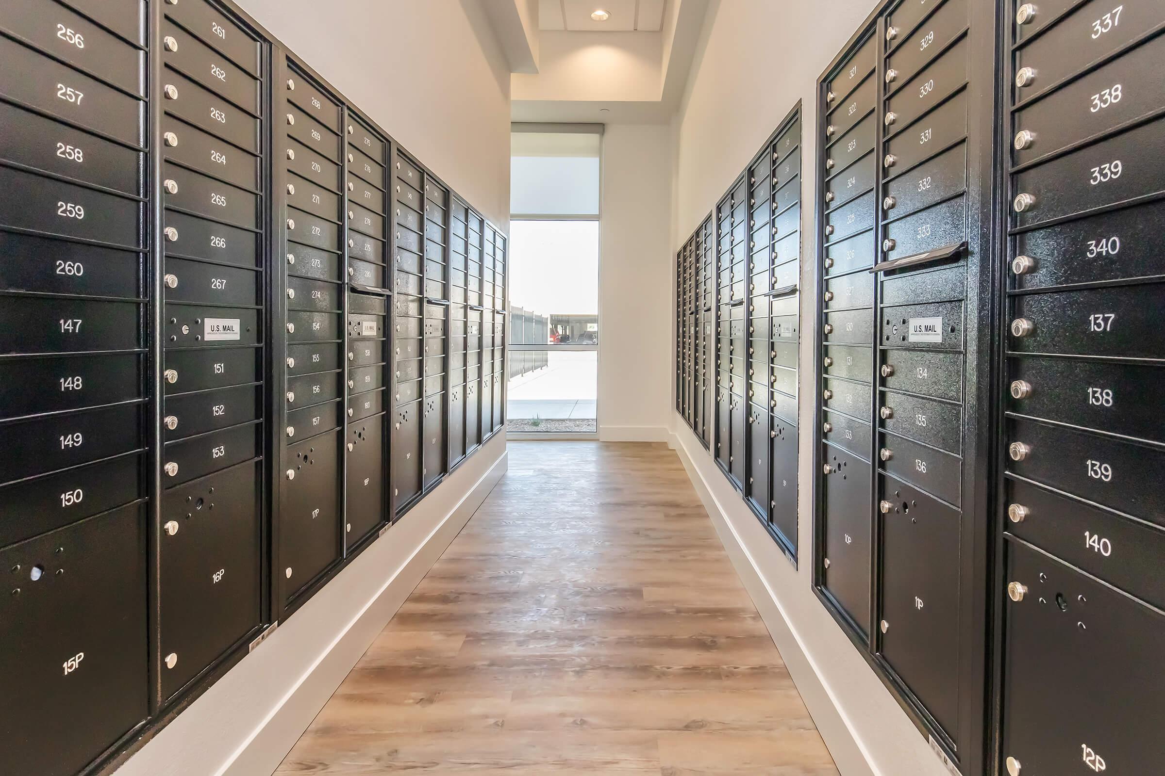 Interior view of a hallway lined with black mailboxes, featuring multiple rows of metal boxes on either side. The floor is wooden, and there's natural light coming in from a window at the end of the corridor. The atmosphere is modern and minimalistic.