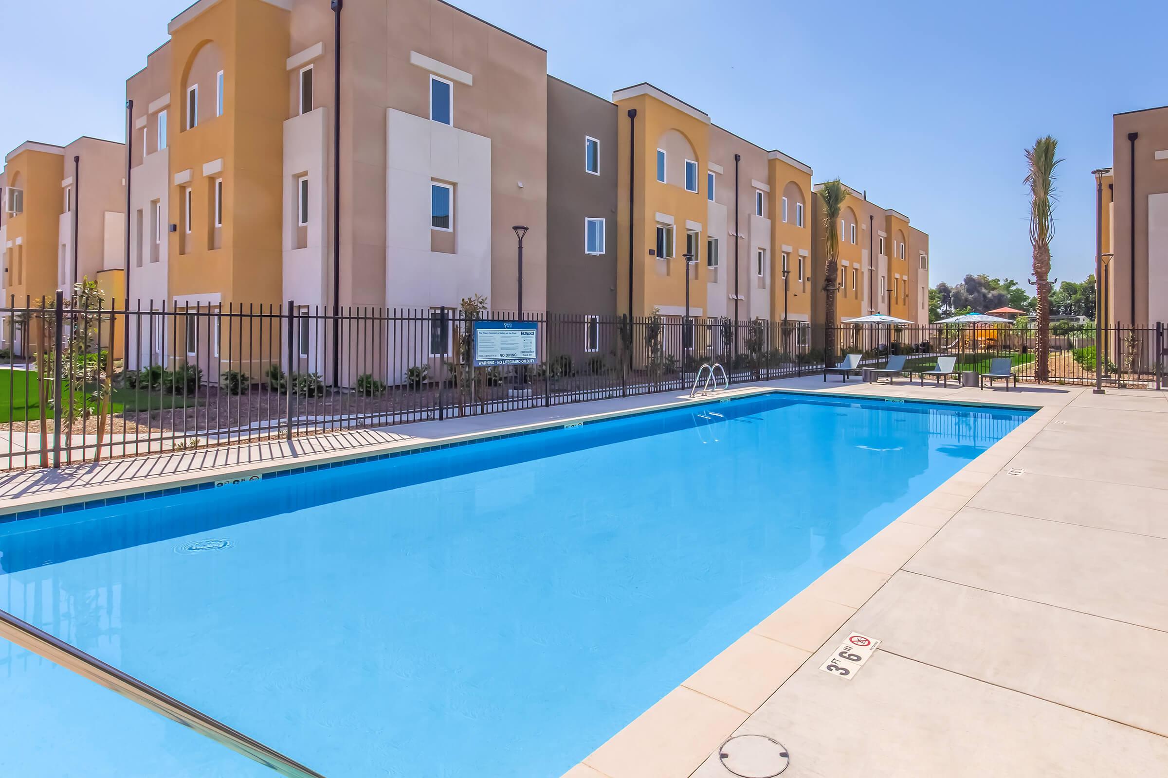 A clear blue swimming pool with a shallow end and pool steps, surrounded by a fence. In the background, there are several modern apartment buildings with beige exteriors. The area is sunny, and there are some lounge chairs and umbrellas nearby, indicating a recreational space. Lush greenery is present around the pool area.